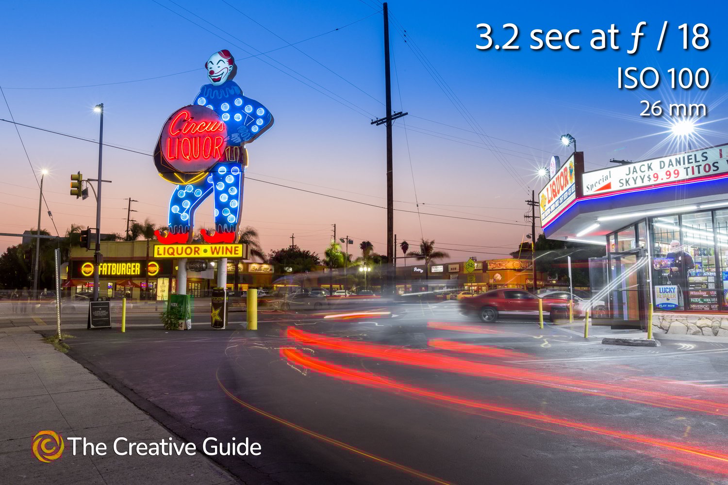 Long exposure night photo of the Circus Liquor neon clown sign with car light trails and surrounding shops, photographed at 3.2 sec f/18 ISO 100, 26 mm, The Creative Guide Photo Gallery.