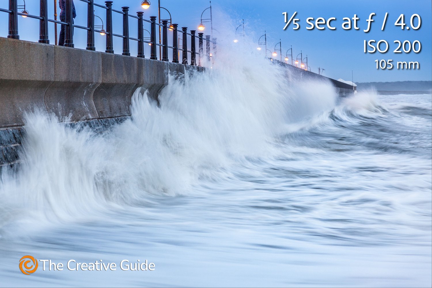 Long exposure of powerful waves crashing against a seawall lined with streetlamps, water motion blurred for effect, photographed at 1/5 sec f/4 ISO 200, 105 mm, The Creative Guide Photo Gallery.