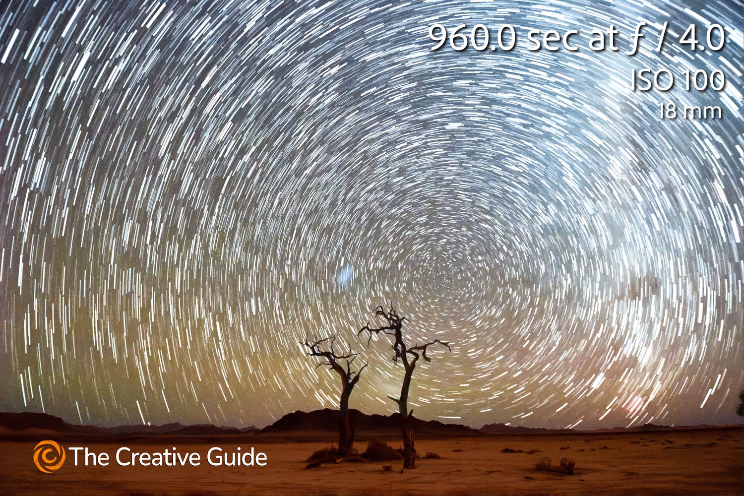 Dramatic star trail photo showing circular patterns in the night sky above two silhouetted desert trees, captured over 9600 seconds f/4 ISO 100, 18 mm, The Creative Guide Photo Gallery.
