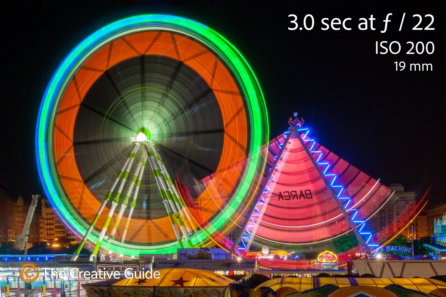 Long exposure photo of a Ferris wheel and fairground ride at night, colorful lights forming circular patterns against the dark sky, photographed at 3 seconds f/22 ISO 200, 19 mm, The Creative Guide Photo Gallery.