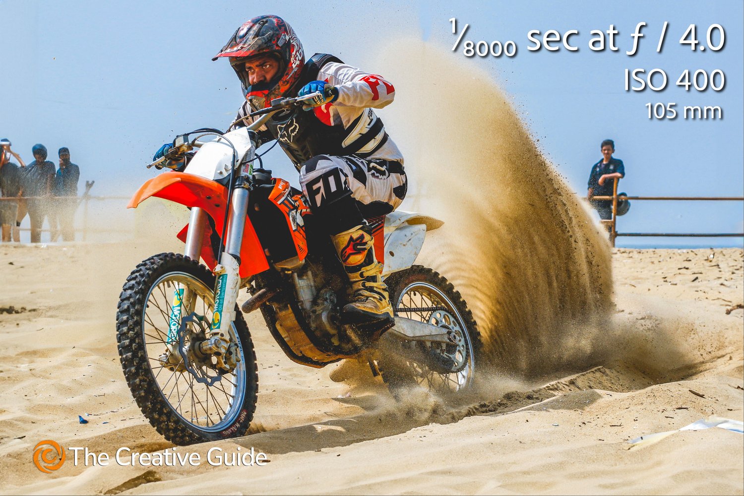 Action shot of a motocross rider leaning into a turn on sandy terrain, kicking up a dramatic spray of sand, photographed at 1/8000 sec f/4 ISO 400, 105 mm, The Creative Guide Photo Gallery.