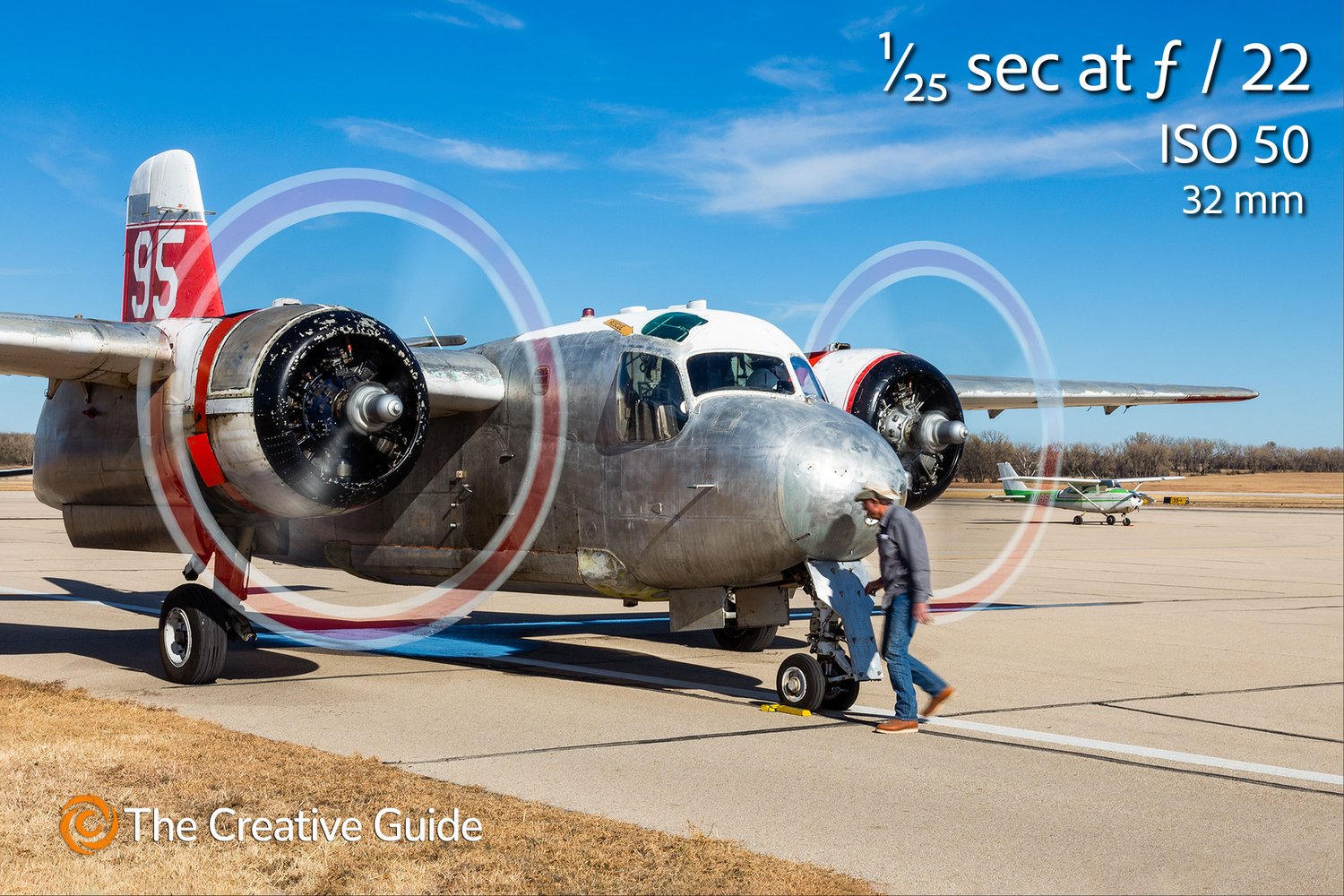 Vintage twin-engine aircraft with propellers spinning on the runway, ground crew member walking past, photographed at 1/25 sec f/22 ISO 50, 32 mm, The Creative Guide Photo Gallery.