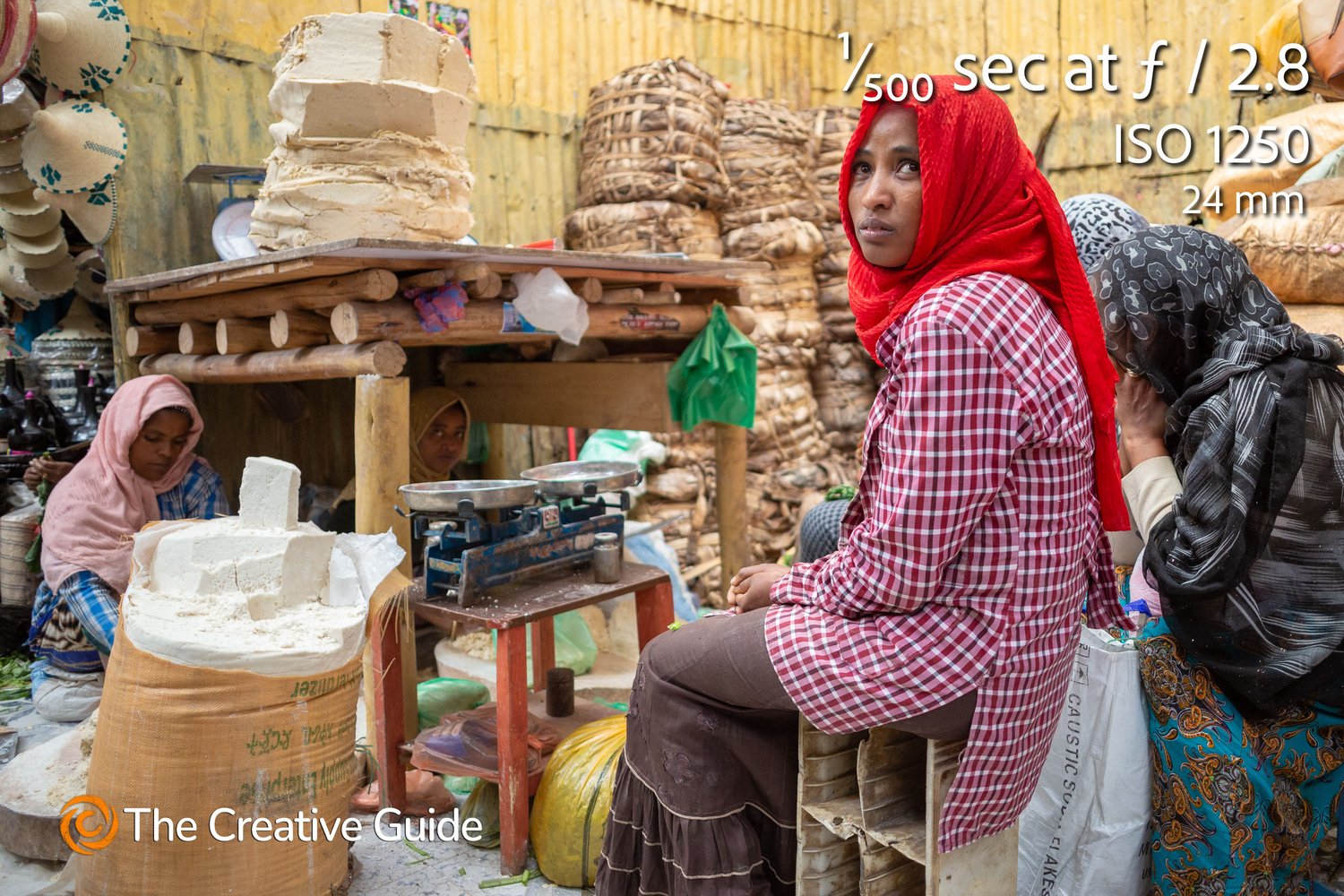 Street market portrait of women beside a stall with large blocks of food and scales, one woman in red headscarf looking directly at camera, photographed at 1/500 sec f/2.8 ISO 1250, 24 mm, The Creative Guide Photo Gallery.