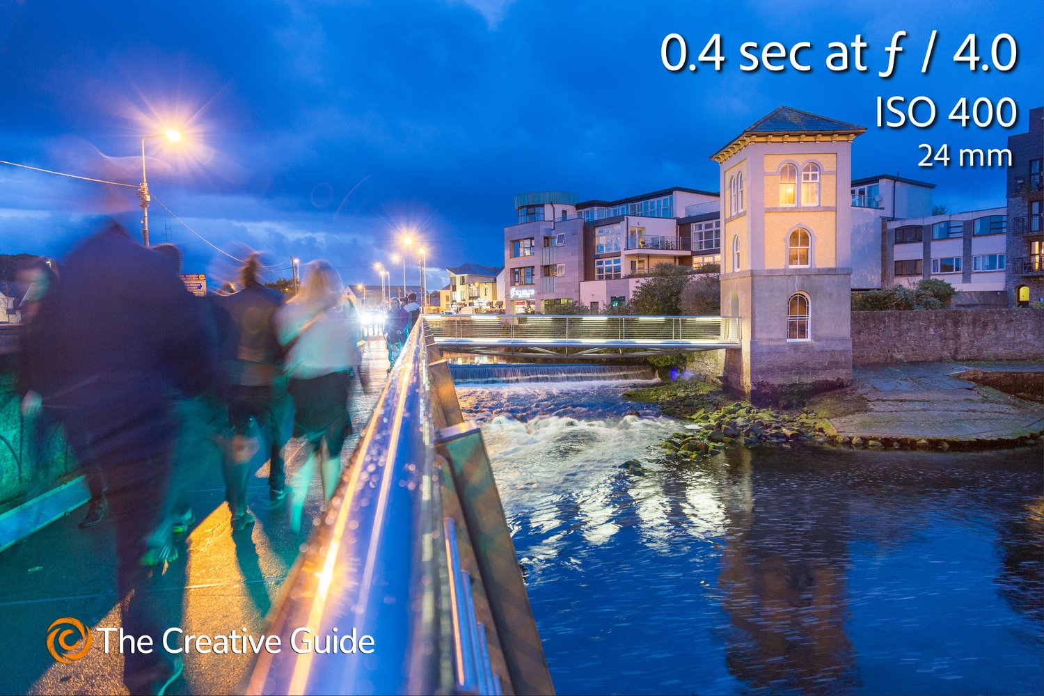 Long exposure street scene at dusk showing blurred figures walking across a bridge, with glowing streetlights and reflections on water, photographed at 0.4 sec f/4 ISO 400, 24 mm, The Creative Guide Photo Gallery.