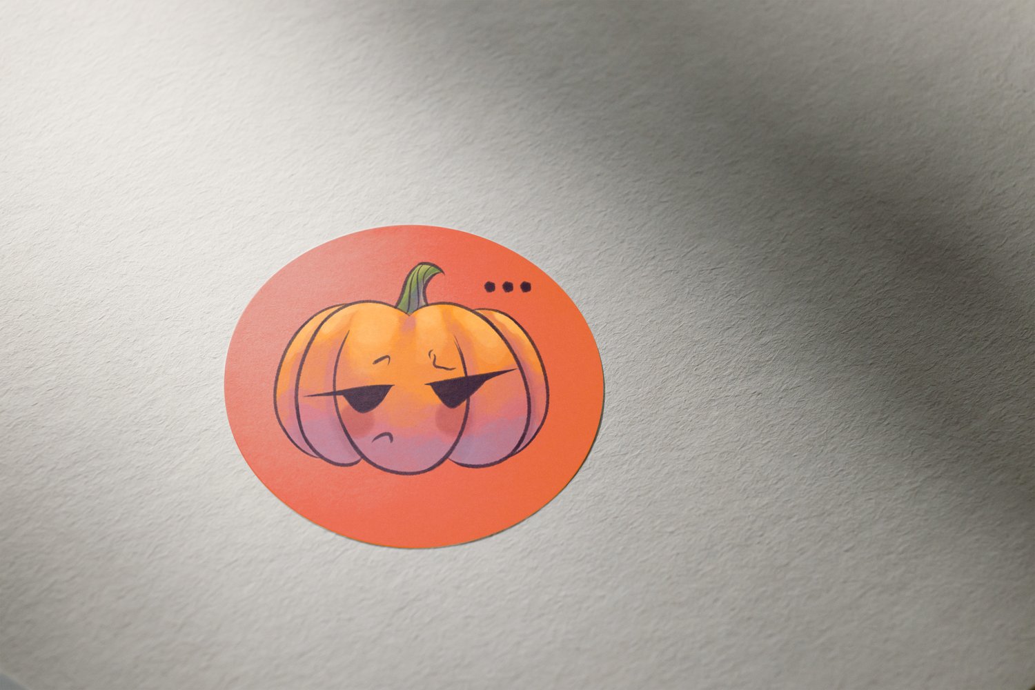 A round, deep orange sticker on a piece of white paper or card. There is a watercolour-inspired illustration of a cartoony jack-o-lantern looking unamused on the sticker. The pumpkin has three dots floating above it.