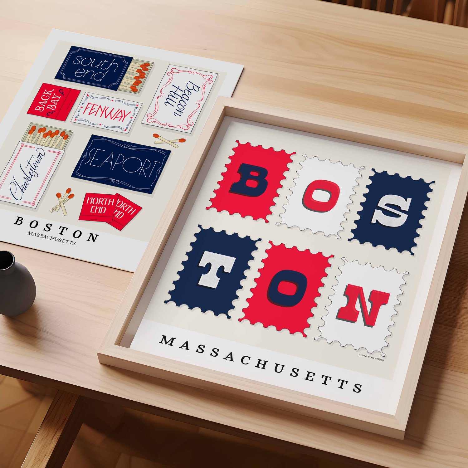 A table with an unframed artwork of Boston matchbox art and a framed artwork of Boston postage stamp art