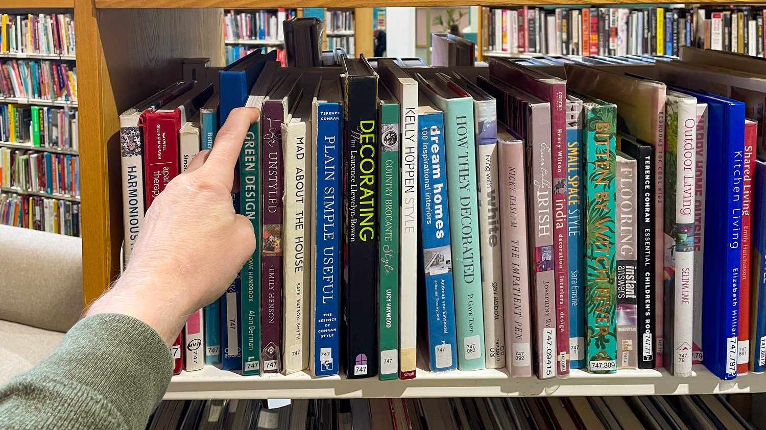 A hand reaching for a random book from a library shelf, surrounded by design and decorating titles, illustrating how The Creative Guide encourages using libraries for creative inspiration.