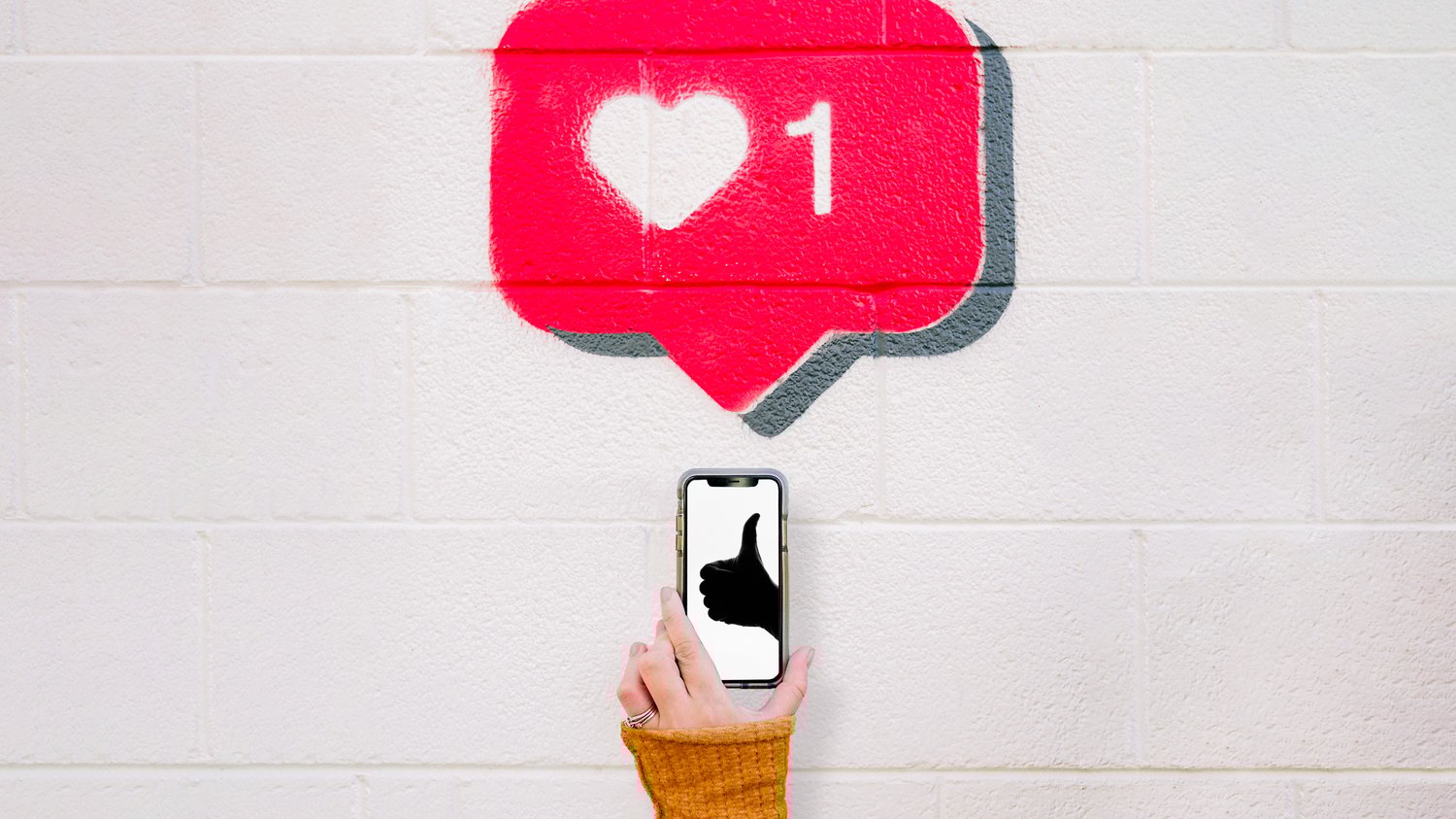 A hand holding a smartphone with a thumbs-up on the screen, raised against a wall painted with a bright red social media like icon, symbolizing the search for approval and how The Creative Guide encourages listening to your own inner voice instead.