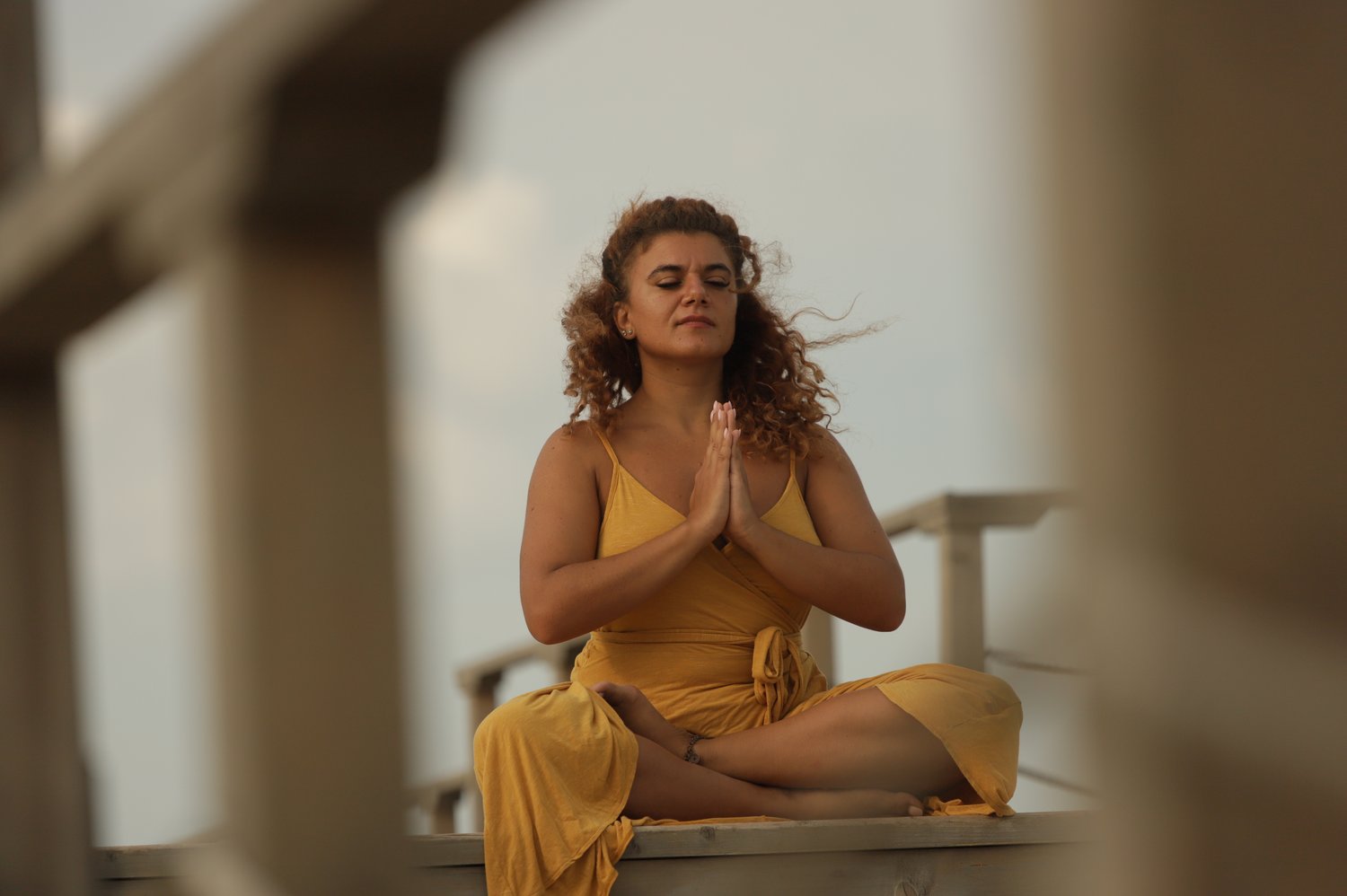 Femeie îmbrăcată în galben meditând în poziție de lotus, cu ochii închiși și palmele împreunate la inimă, practicând yoga și conectare spirituală în aer liber la apus