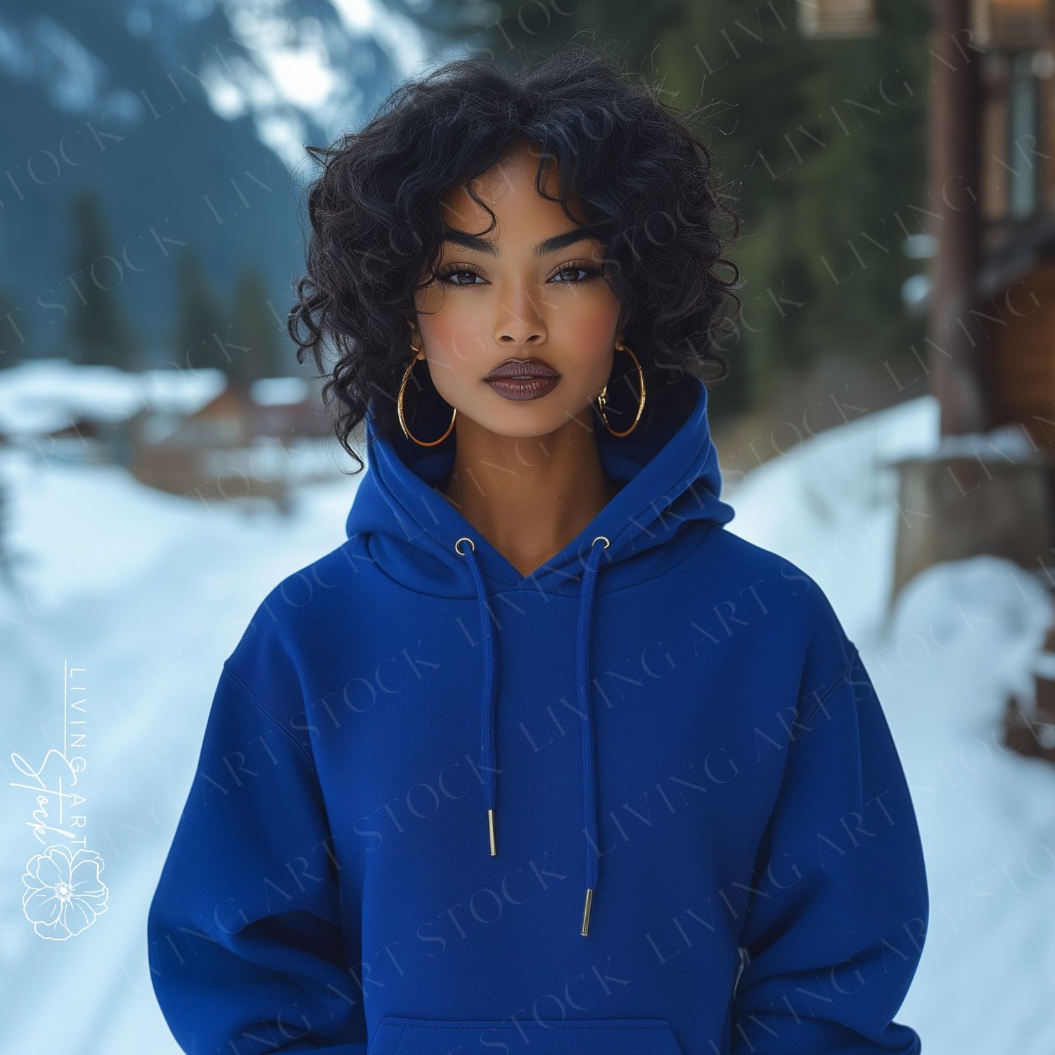 A beautiful woman with striking, dark, curly hair and bold makeup is captured in a close-up portrait outdoors, set against a snowy, mountainous backdrop. She is wearing a vibrant, royal blue hoodie with a hood drawn up slightly, and large gold hoop earrin