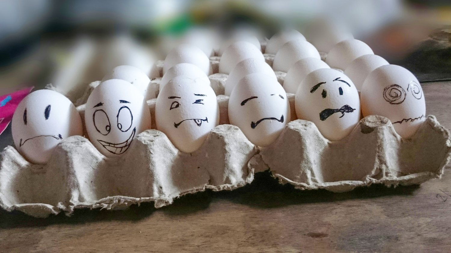 A carton of white eggs with expressive faces drawn on them, each showing a different emotion, symbolizing individuality, perception, and group dynamics in creative thinking for The Creative Guide.