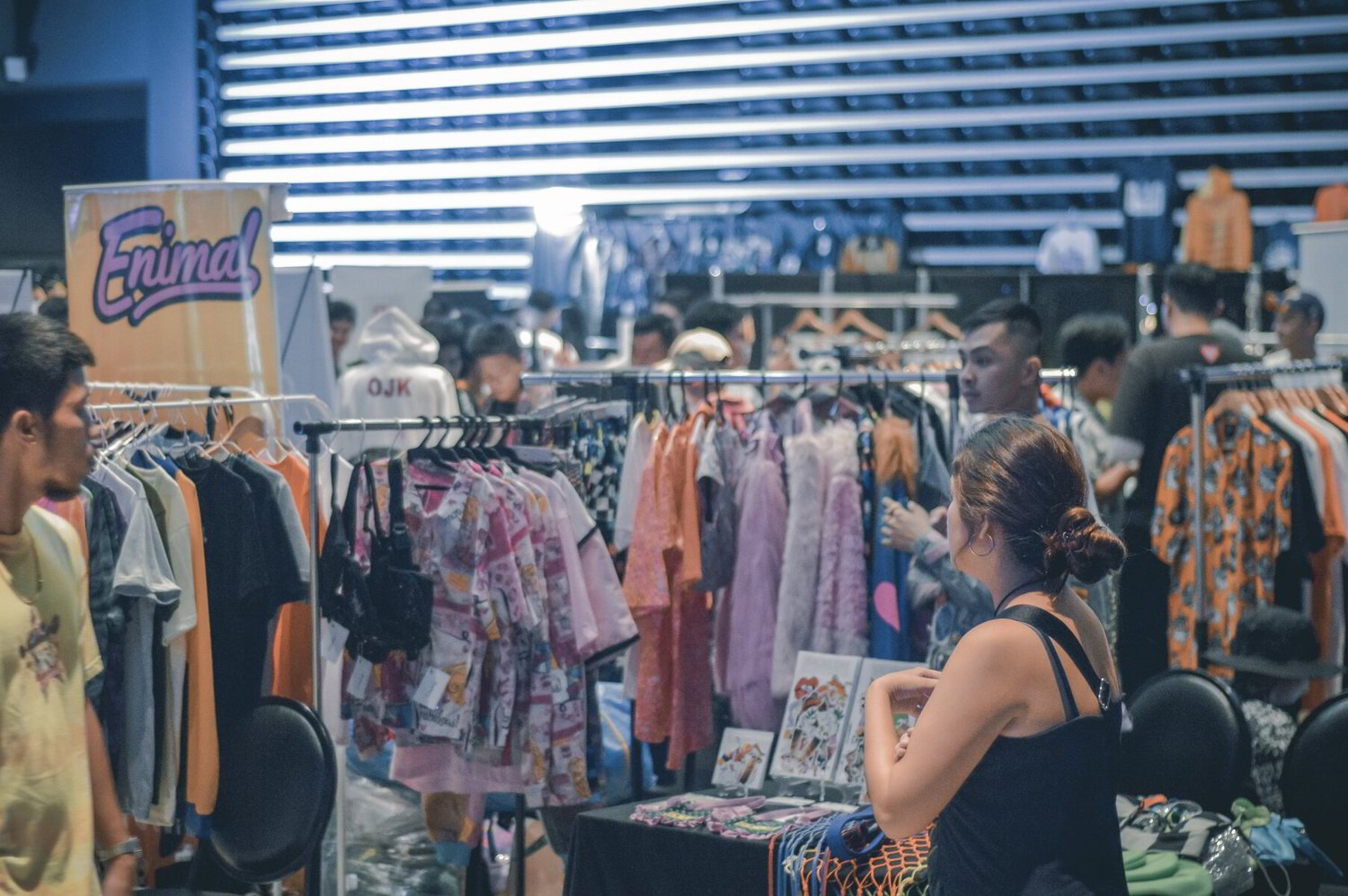 Indoor market scene with colorful clothing racks and busy shoppers. A woman stands near a clothing stall, creating a lively and bustling atmosphere.