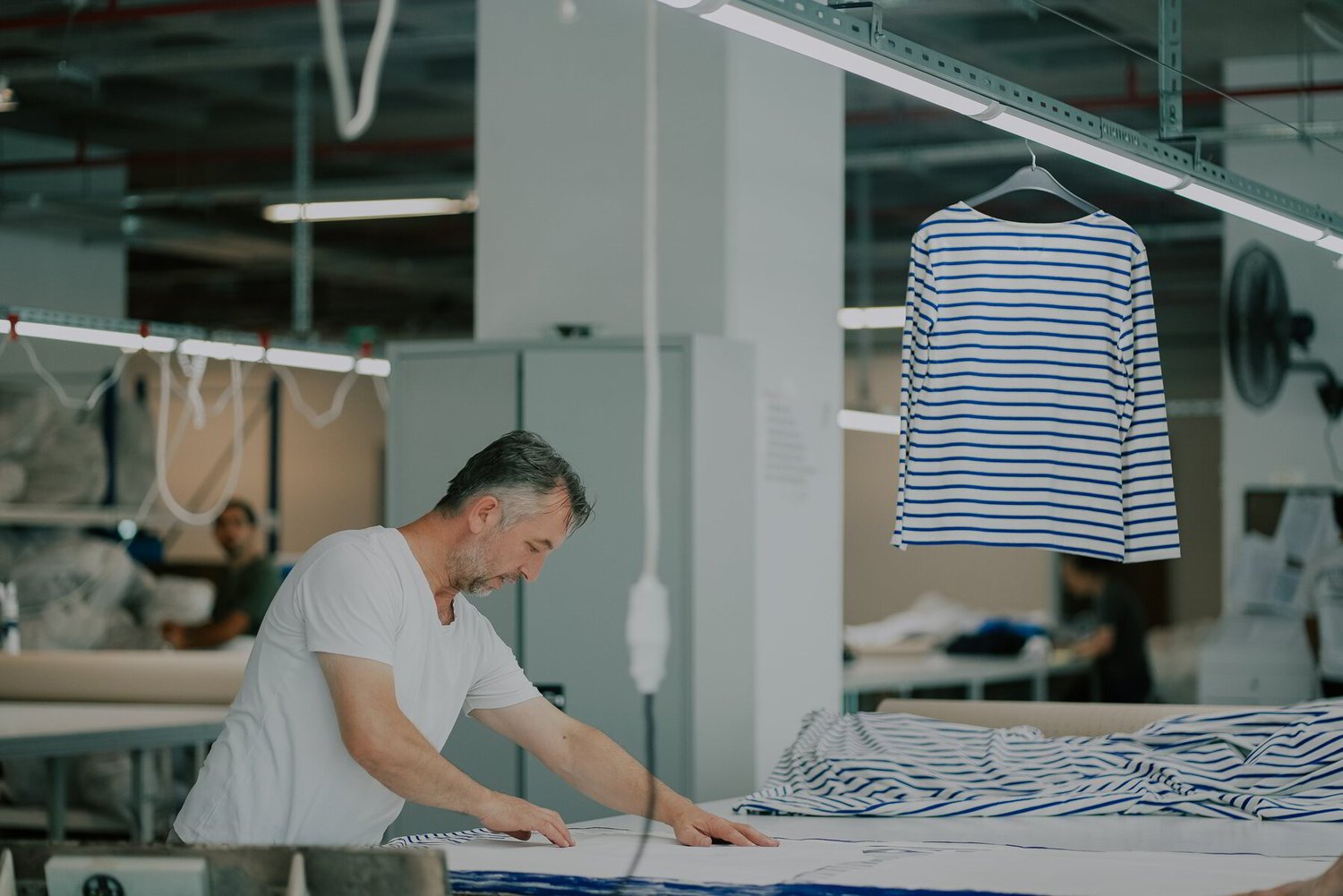 Man in a white shirt works at a textile factory, examining striped fabric. A blue and white striped shirt hangs nearby, conveying focus and craftsmanship.