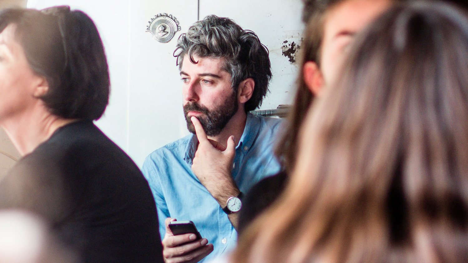 A man in a blue shirt holds a phone and rests his hand on his chin while listening during a group discussion, reflecting the thought and attentiveness explored in The Creative Guide’s “Sunday Reflection.”
