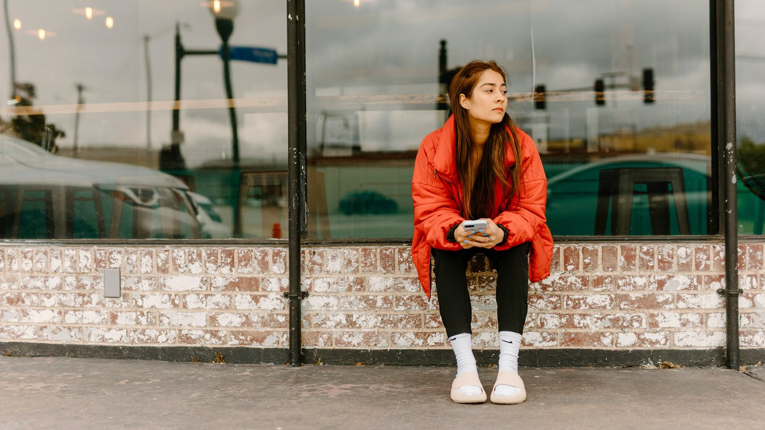 A person wearing a bright red jacket sits on a low brick ledge outside a café, holding a phone and looking sideways toward the street, suggesting a moment of pause and shifted attention in The Creative Guide’s “Thinking Differently.”