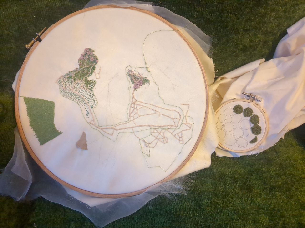One large and one small embroidery hoops on a green fabric background. The hoops contain off-white fabric. The larger one has the rough shapes of an aerial view depicted in a variety of stitches. The smaller hoop contains circles of textured knot stitches