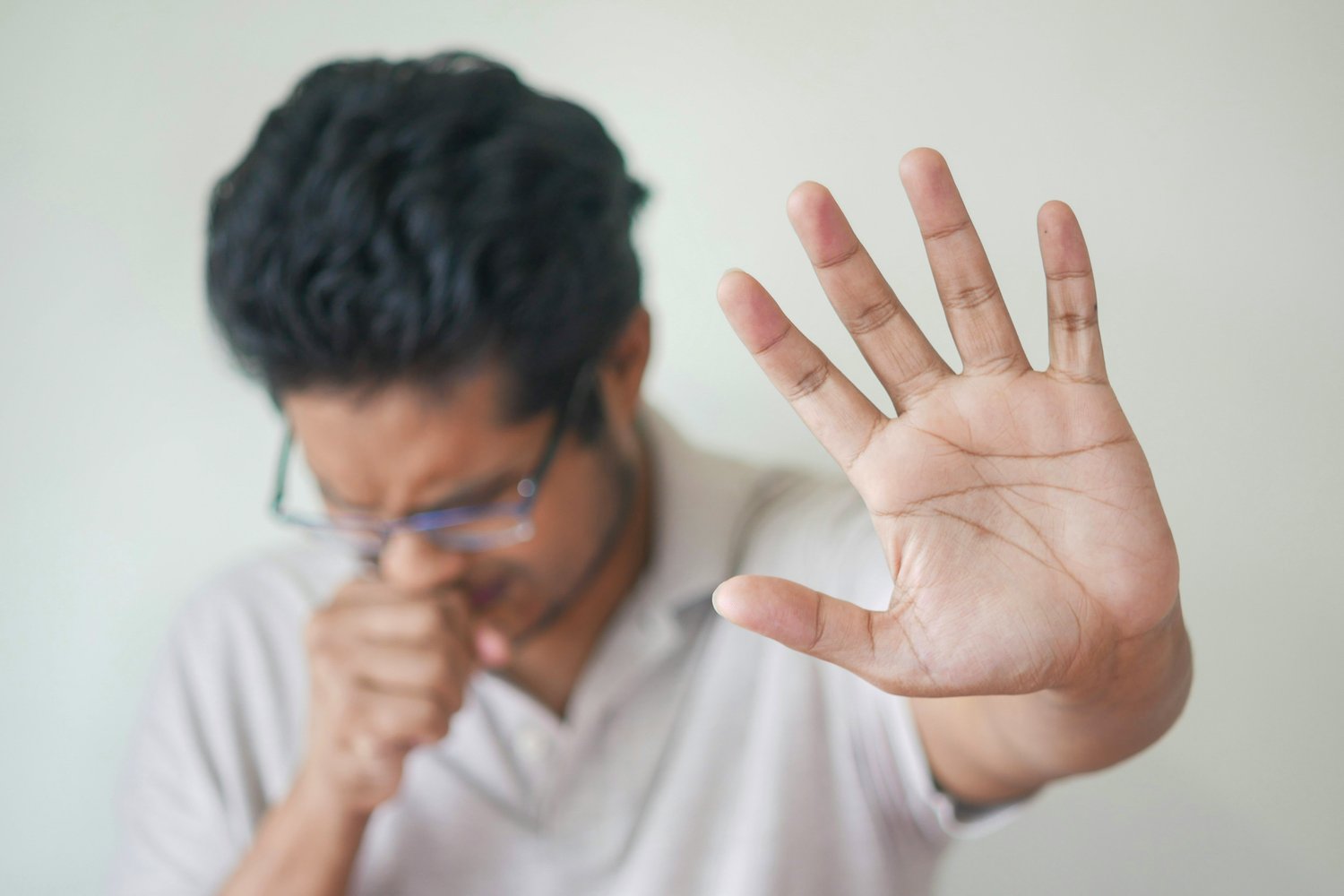 A man wearing glasses and a light shirt sneezes into his hand while raising his other hand in a stopping gesture, his expression tense and eyes closed, visually capturing the sudden interruption described in The Creative Guide’s “Inner Work.”