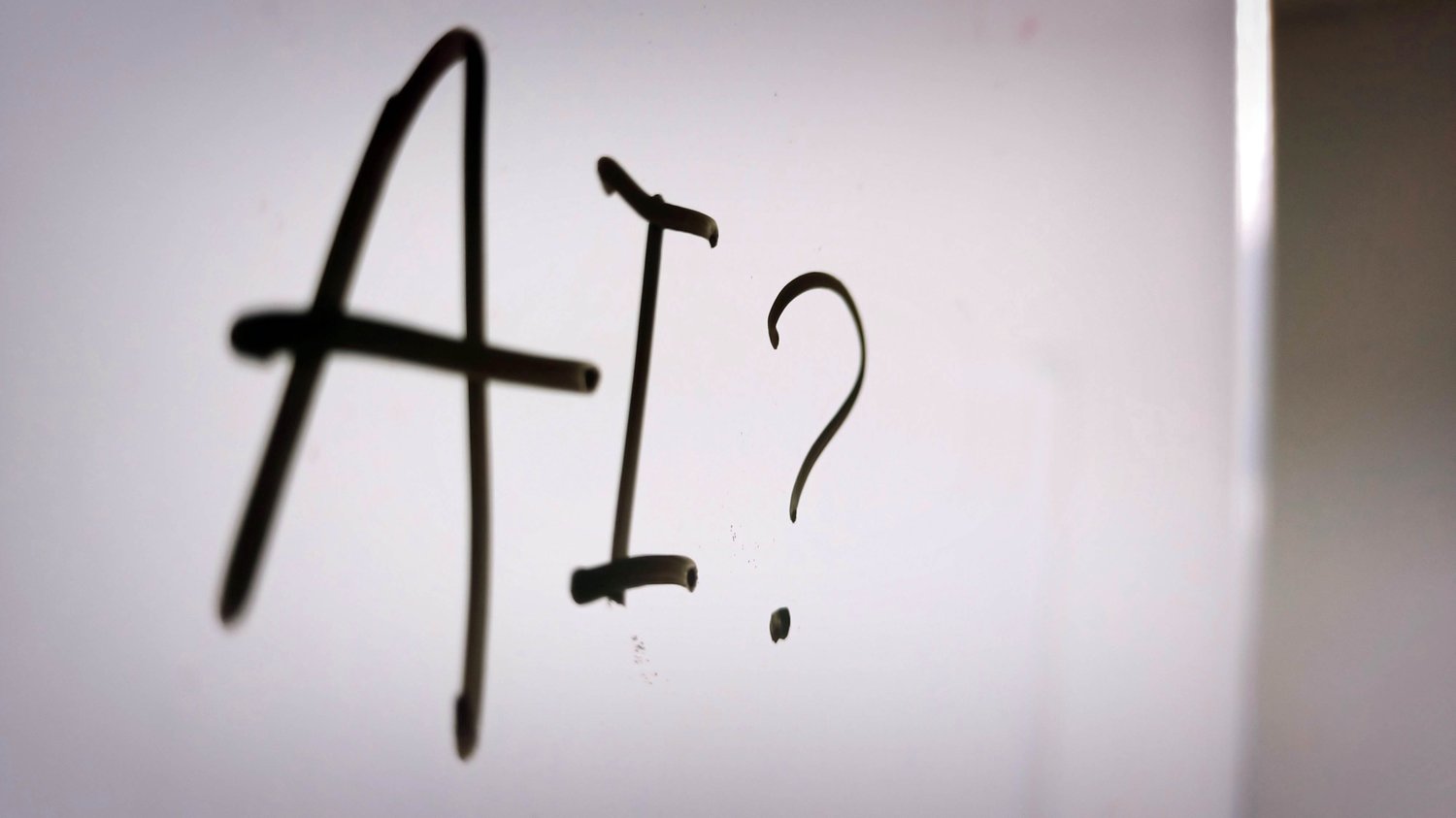 The letters AI followed by a question mark are written in dark marker on a smooth reflective surface that could be glass or a whiteboard, with soft natural light coming from the right and fading into shadow on the left. The writing is slightly blurred at 