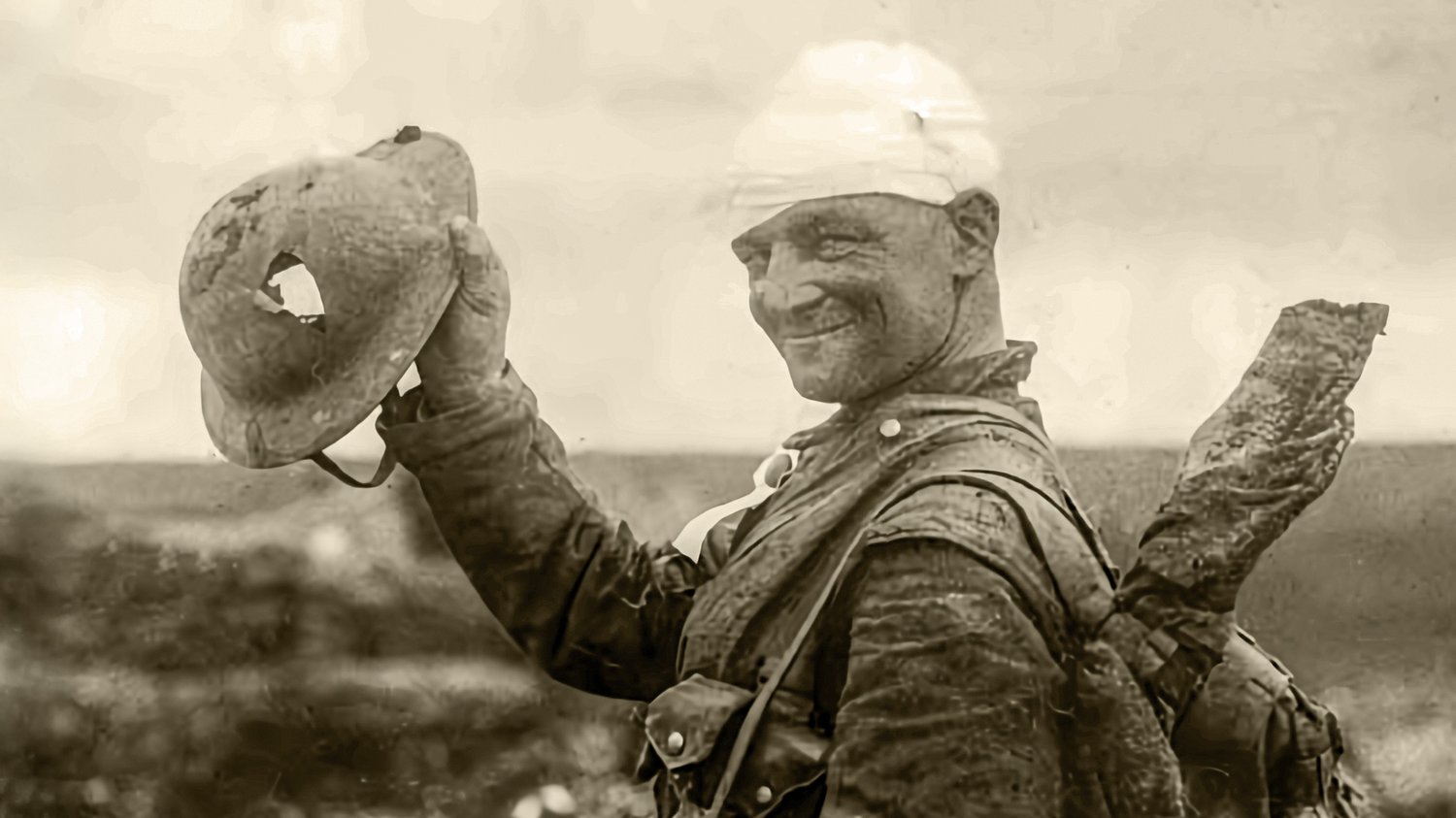 A sepia-toned photograph shows a World War 1 soldier with a bandaged head raising a shredded steel helmet toward the camera, his face marked with dirt yet holding a small smile, suggesting survival and relief after the helmet saved him, matching The Creat