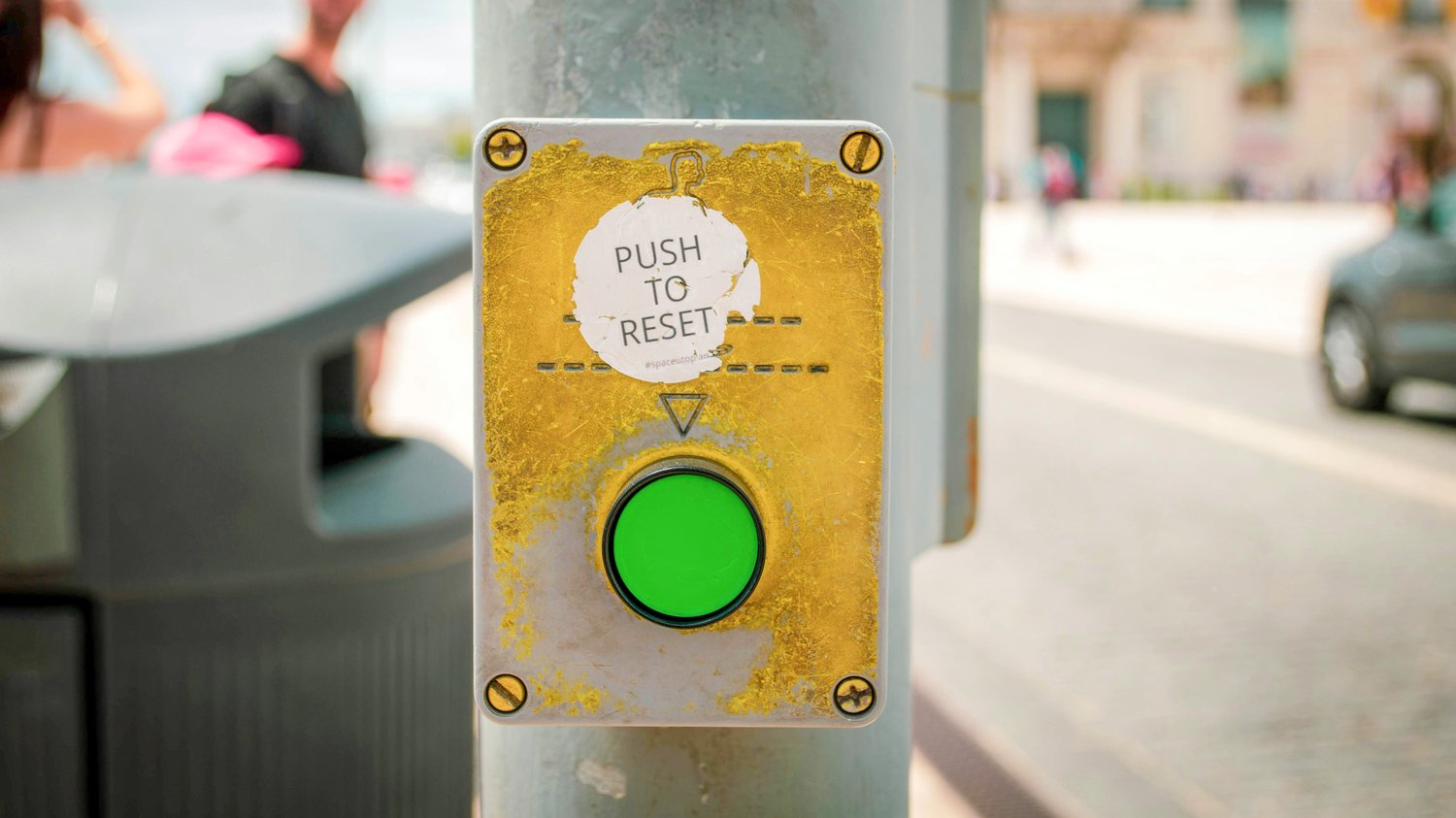 A close-up photograph of a weathered pedestrian button on a city pole with a bright green light, above it a torn sticker that reads “PUSH TO RESET,” while people and cars move slightly out of focus in the background, matching The Creative Guide’s idea of 