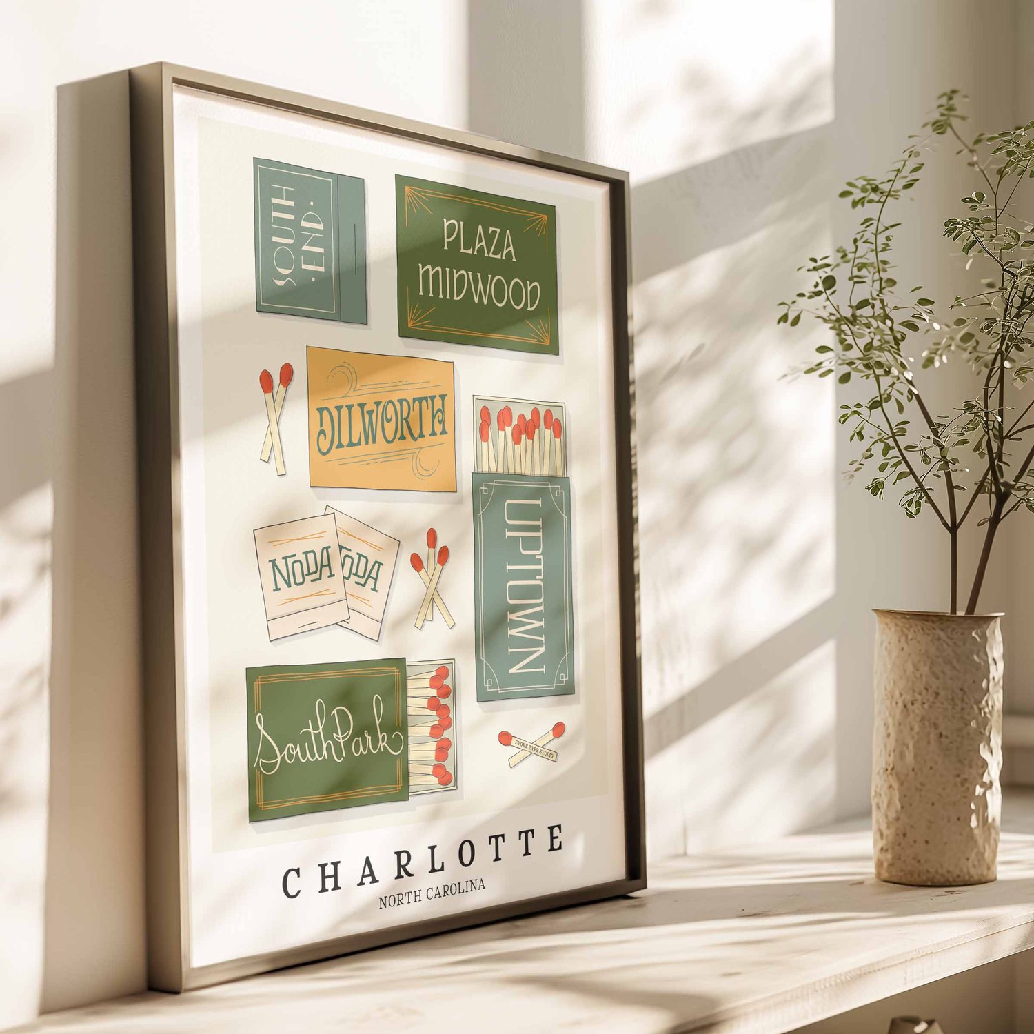 A framed picture of Charlotte, North Carolina neighborhoods drawn in various lettering styles on matchboxes. Artwork has a blue, green and yellow color scheme.