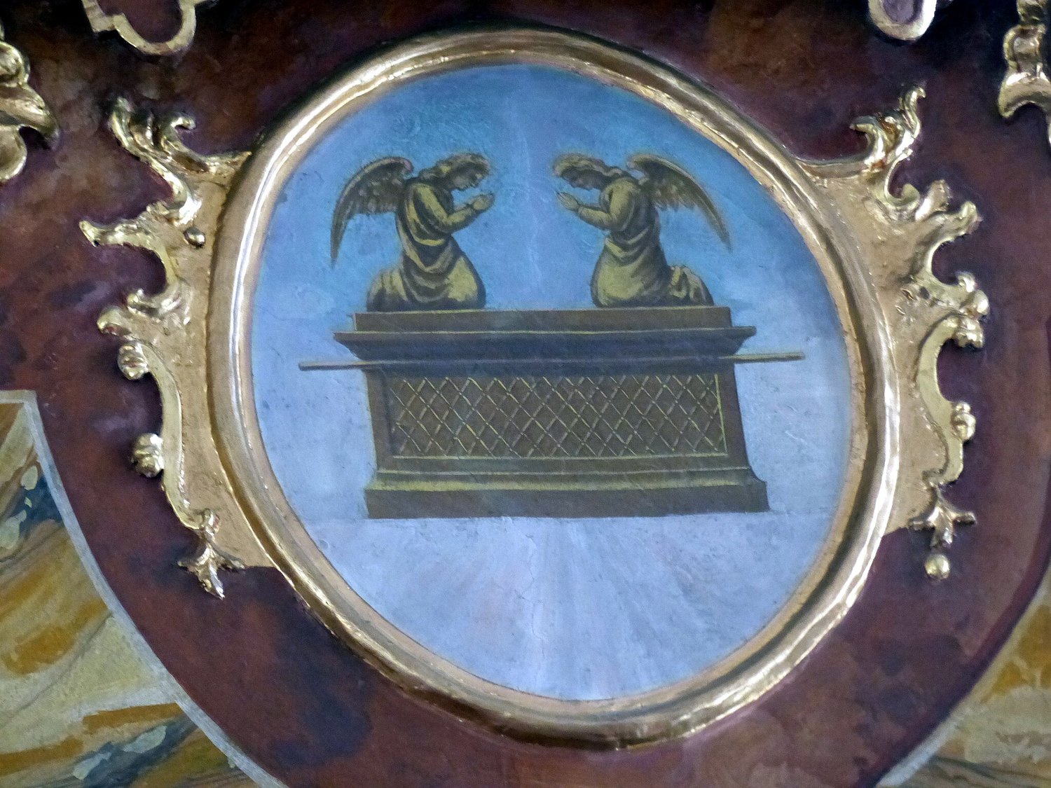 A photograph of a painted ceiling depicting the Ark of the Covenant - a box topped by two cherubim (angels) facing each other and praying. The Ark is in a blue circle surrounded by silver decoration. It is above the Altar of the Holy Family in Saint Barth