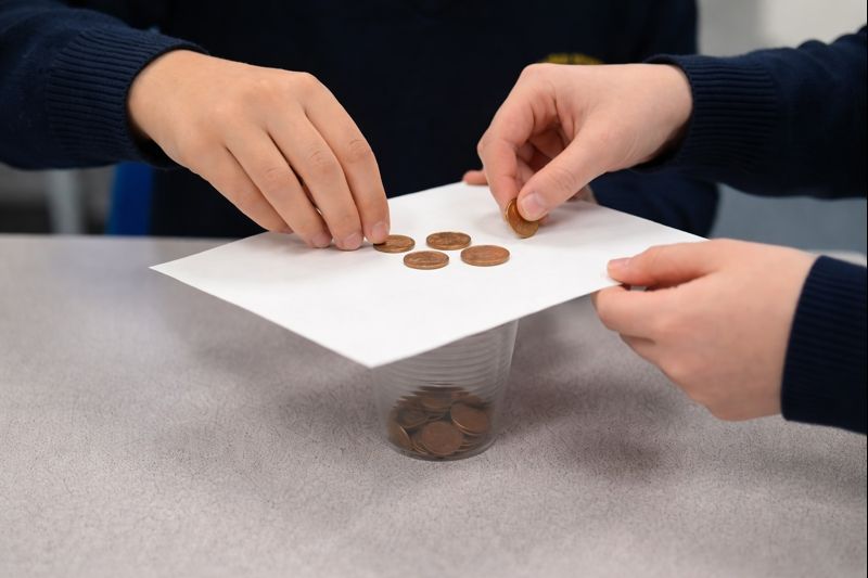 Close-up of two students’ hands placing copper coins onto a sheet of white paper balanced on top of a clear plastic cup during a classroom science experiment.