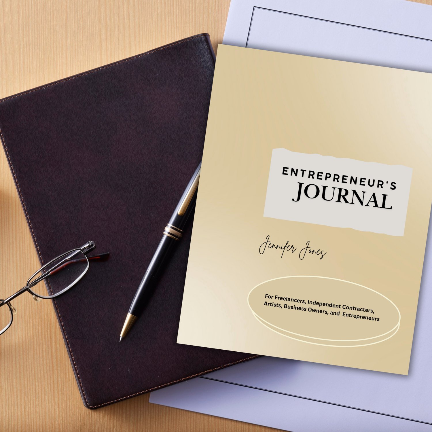 This image depicts the book cover of  the digital printable notebook the "Entrepreneur's Journal" by Jennifer Jones "for freelancers, independent contractors, artists, business owners, and entrepreneurs," as cited on the bottom of the gold colored  cover. The journal is set on top of a desk arrayed with  a black notebook, eyeglass, pen, and paper.
