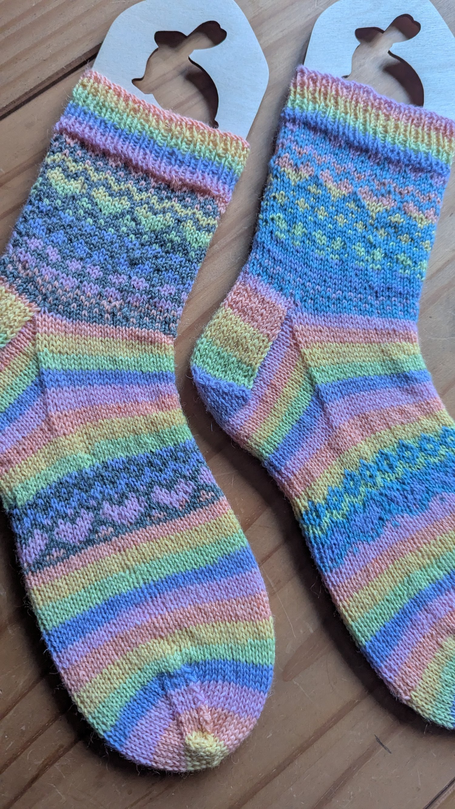 a pair of knitted socks on blockers in muted rainbow yarn with colour work in heart shape patterns