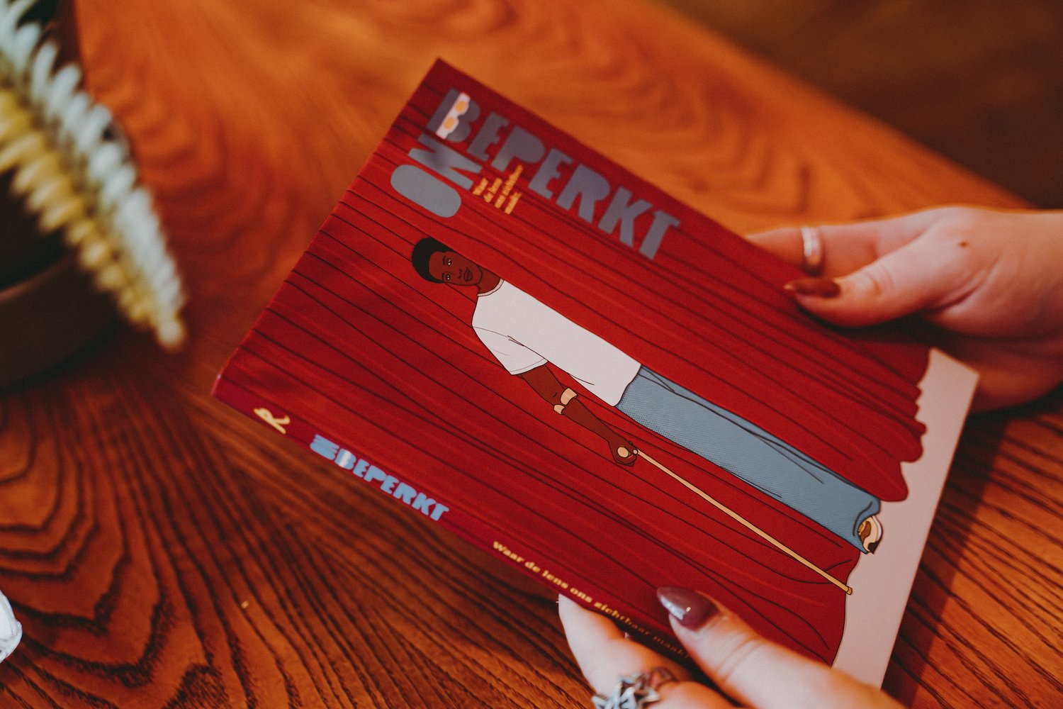Boek met de titel “Onbeperkt” wordt door twee handen vast gehouden boven een houten tafel. De omslag van het boek toont een illustratie van een persoon voor rode gordijnen met een gele wandelstok.