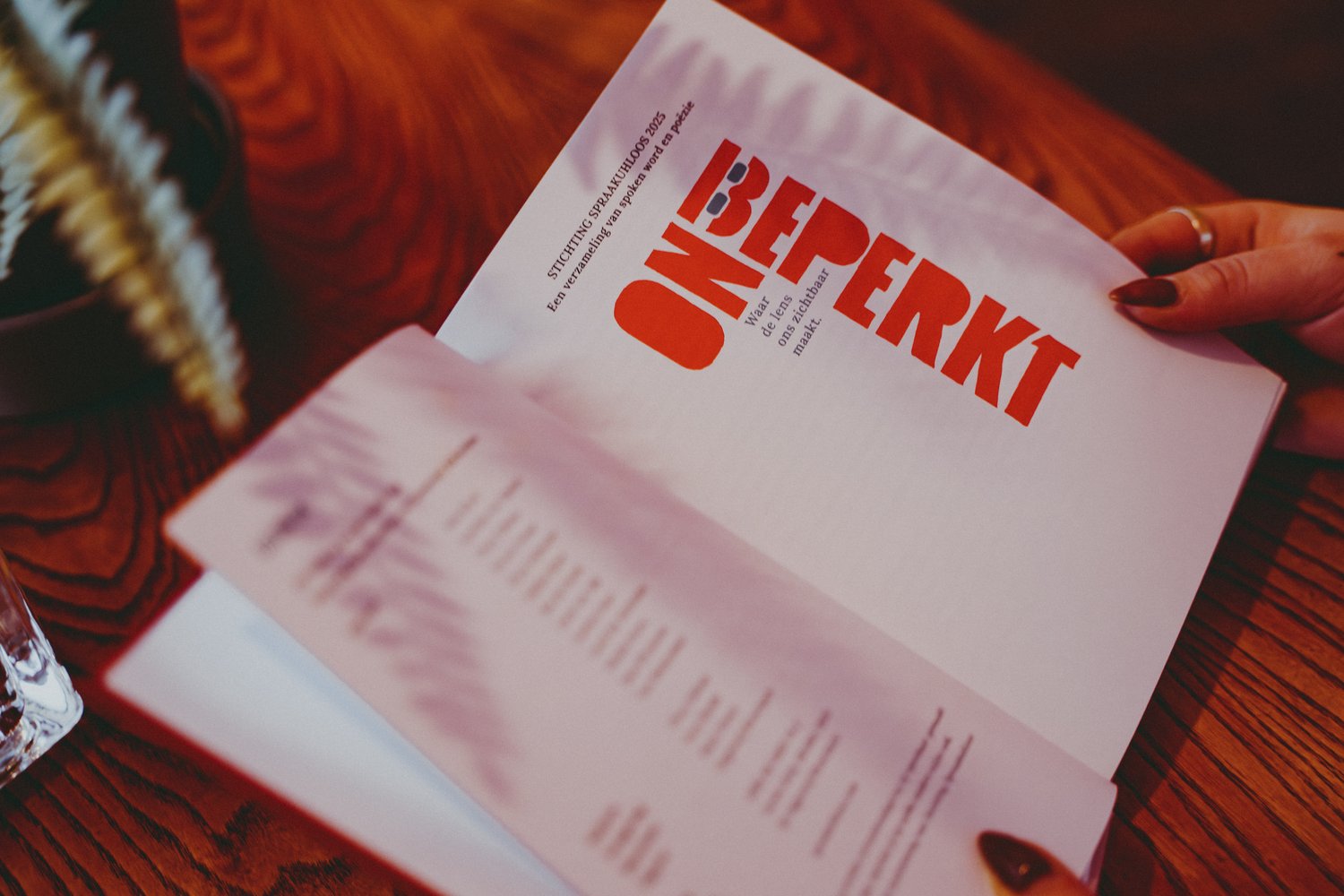 Open boek dat wordt vastgehouden boven een houten tafel. Op de rechterpagina staat de titel “Onbeperkt” met kleinere tekst eronder. Op de linkerpagina staat een colofon pagina.