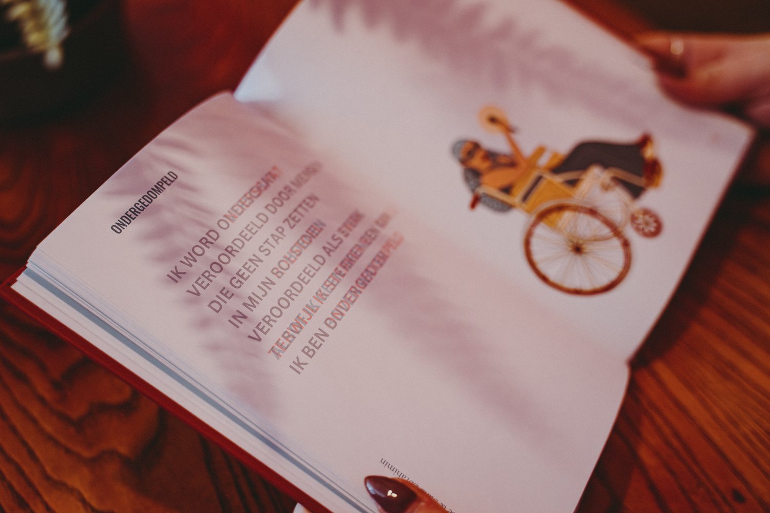 Open boek op een houten tafel. Op de linkerpagina staat een anaglyf gedicht in het Nederlands. Op de rechterpagina staat een illustratie van een vrouw in een rolstoel, ze houdt een microfoon vast in haar hand.