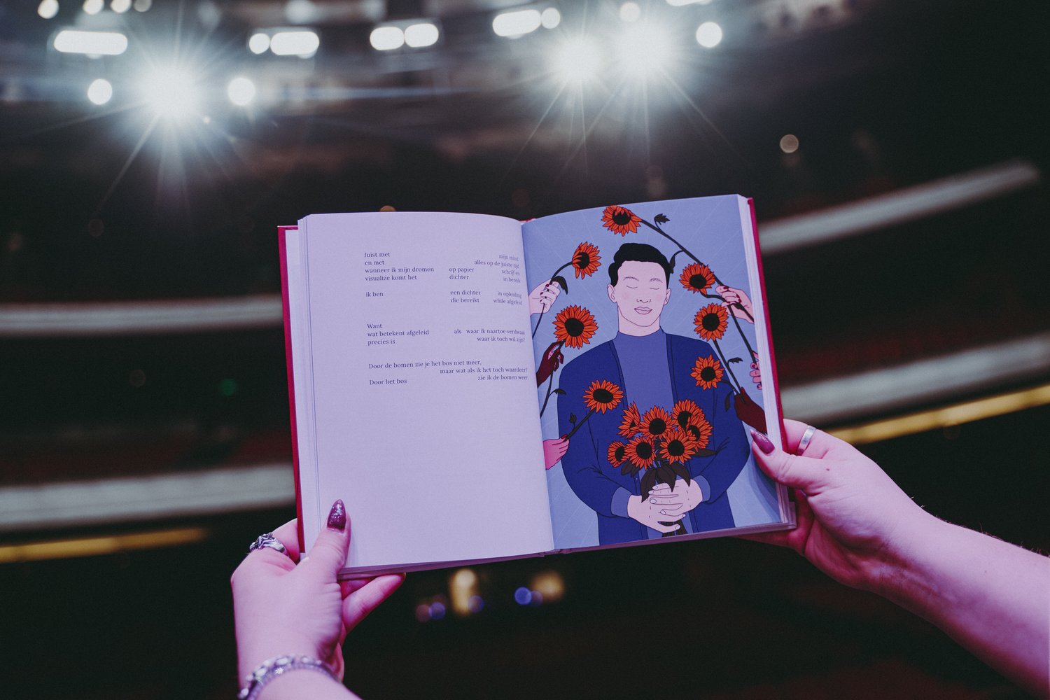 Foto van twee handen die een open boek vasthouden in een theaterzaal. Op de achtergrond zijn felle podiumlichten zichtbaar. Op de linkerpagina staat een Nederlandstalig gedicht in zwarte tekst op een witte pagina. Op de rechterpagina staat een illustratie van een persoon met gesloten ogen die een boeket zonnebloemen vasthoudt. Rond de geïllustreerde persoon zijn meerdere handen te zien die zonnebloemen aanbieden.