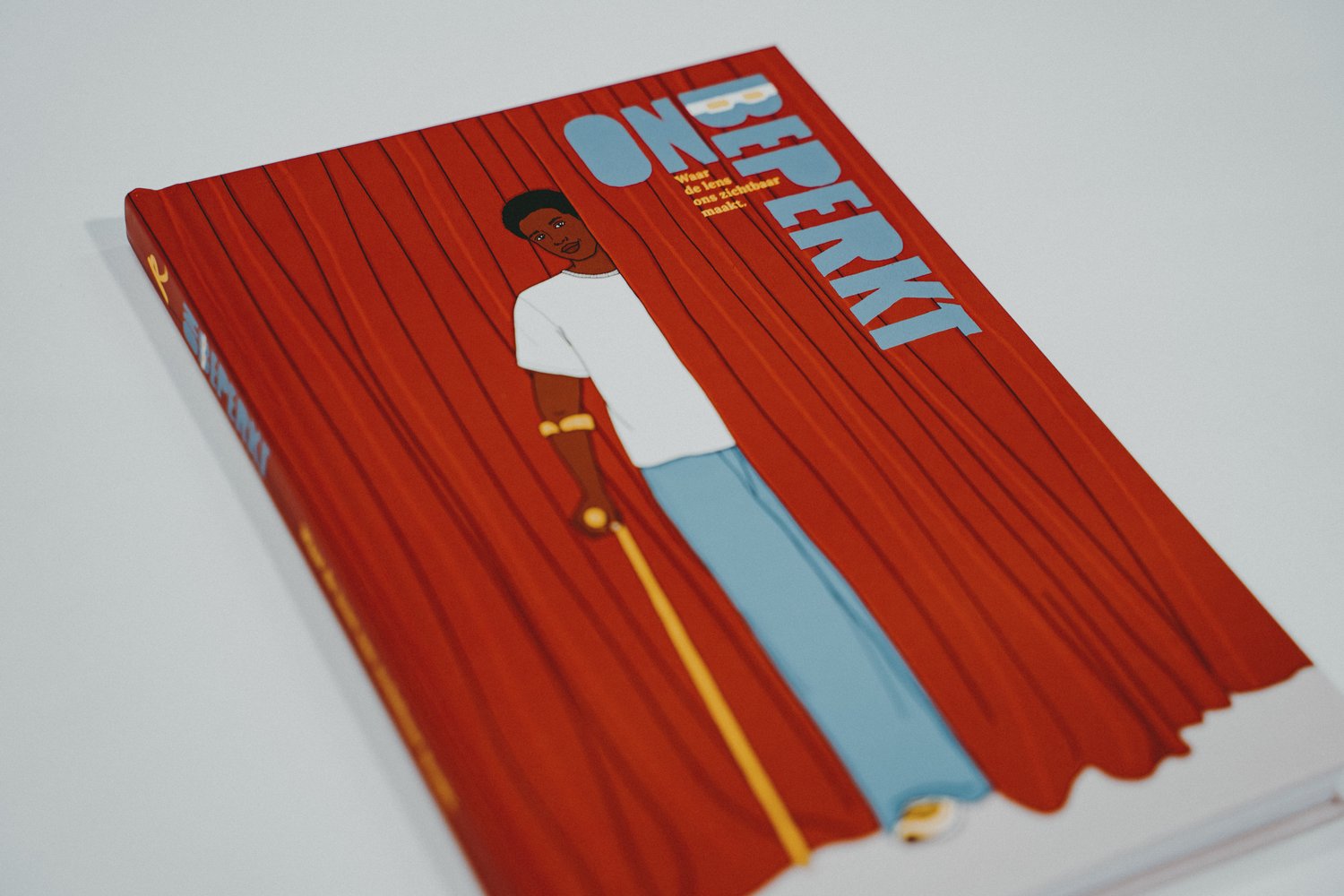 Schuin gefotografeerd boek met de omslagtitel “Onbeperkt” tegen een lichte achtergrond. De illustratie op de omslag toont een persoon voor rode gordijnen met een gele wandelstok.