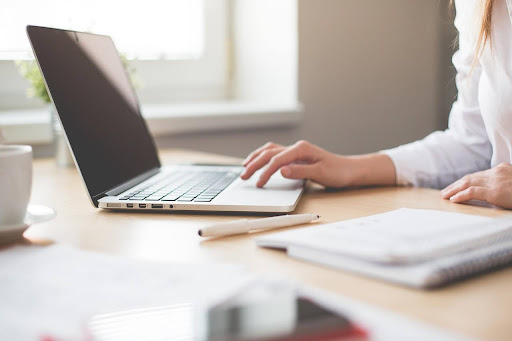 a light wood desk with a laptop on it and a persons arm/hand touching the laptop (the rest of the person is out of shot) along with a notepad, mug and pen.
