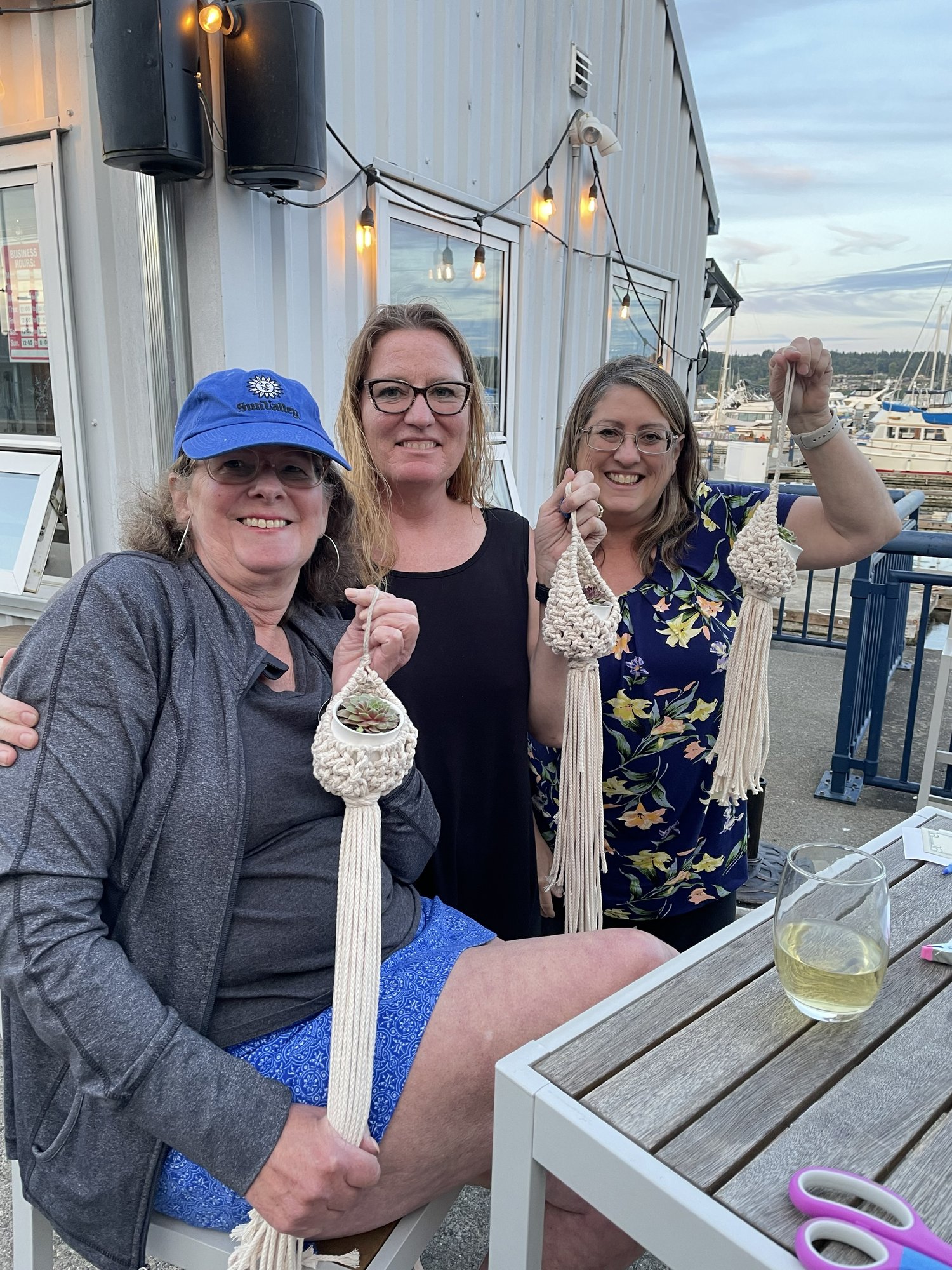 Group of participants smiling and displaying their completed macramé plant hangers during an outdoor workshop.