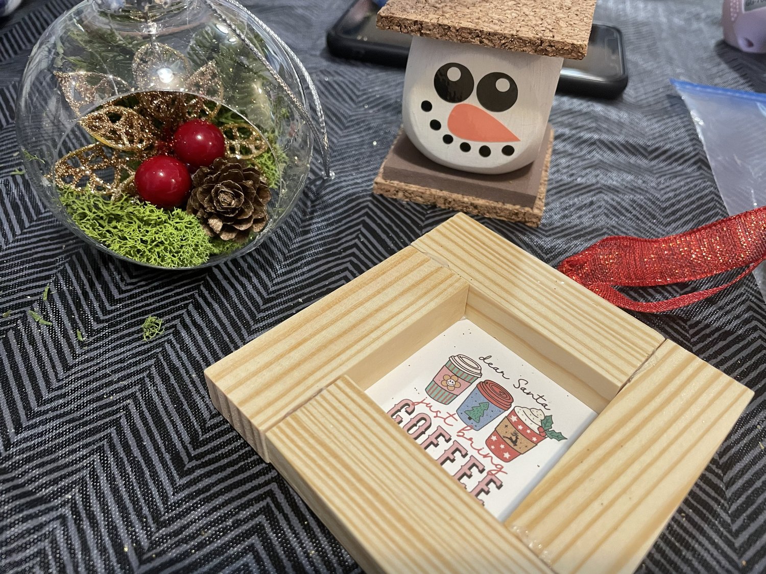 Tabletop display of craft workshop projects including a moss‑filled ornament with pinecones and berries, a framed coffee‑themed mini print, and a small handmade snowman decoration.