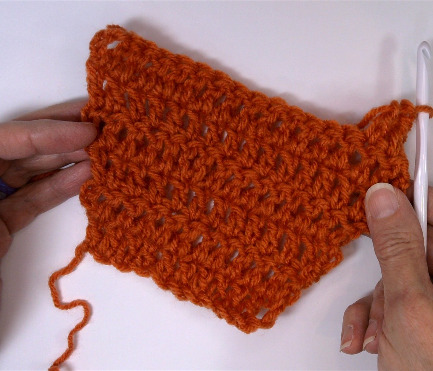 Close-up of a crochet swatch getting wider at the edges, demonstrating extra stitches in rows with a white hook and hands holding the fabric
