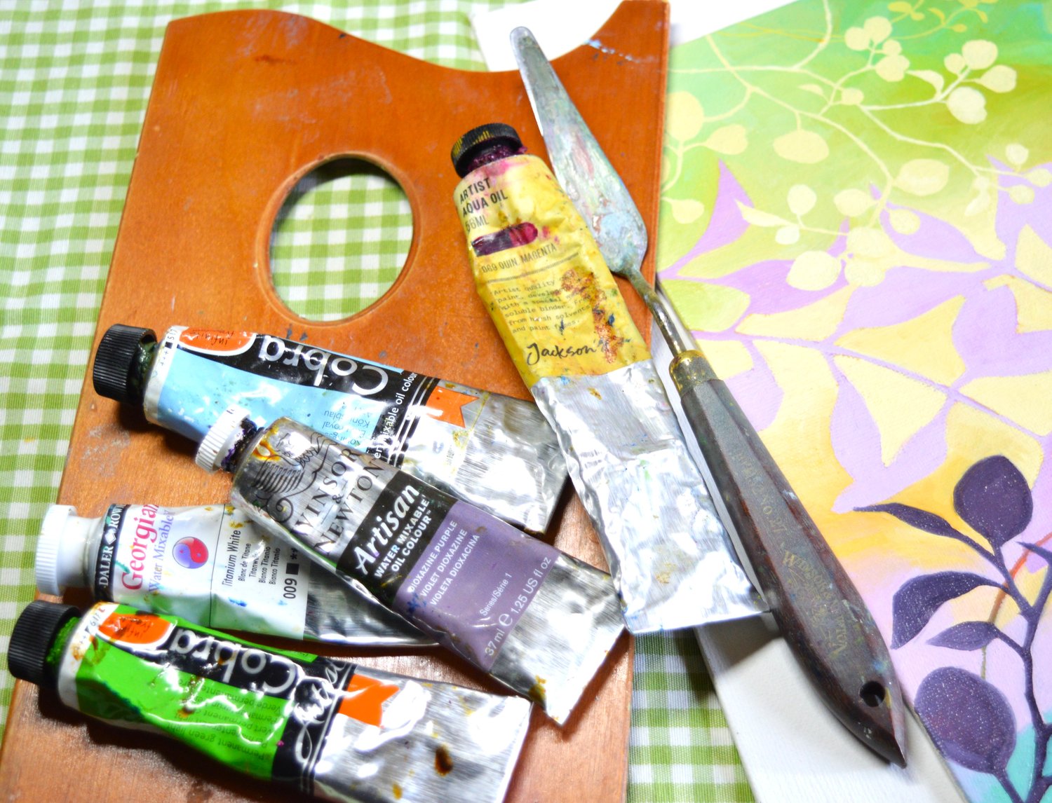tubes of oil paints on a wooden palette with painting knife and a decorative modern floral oil painting all on a light green checkered cloth