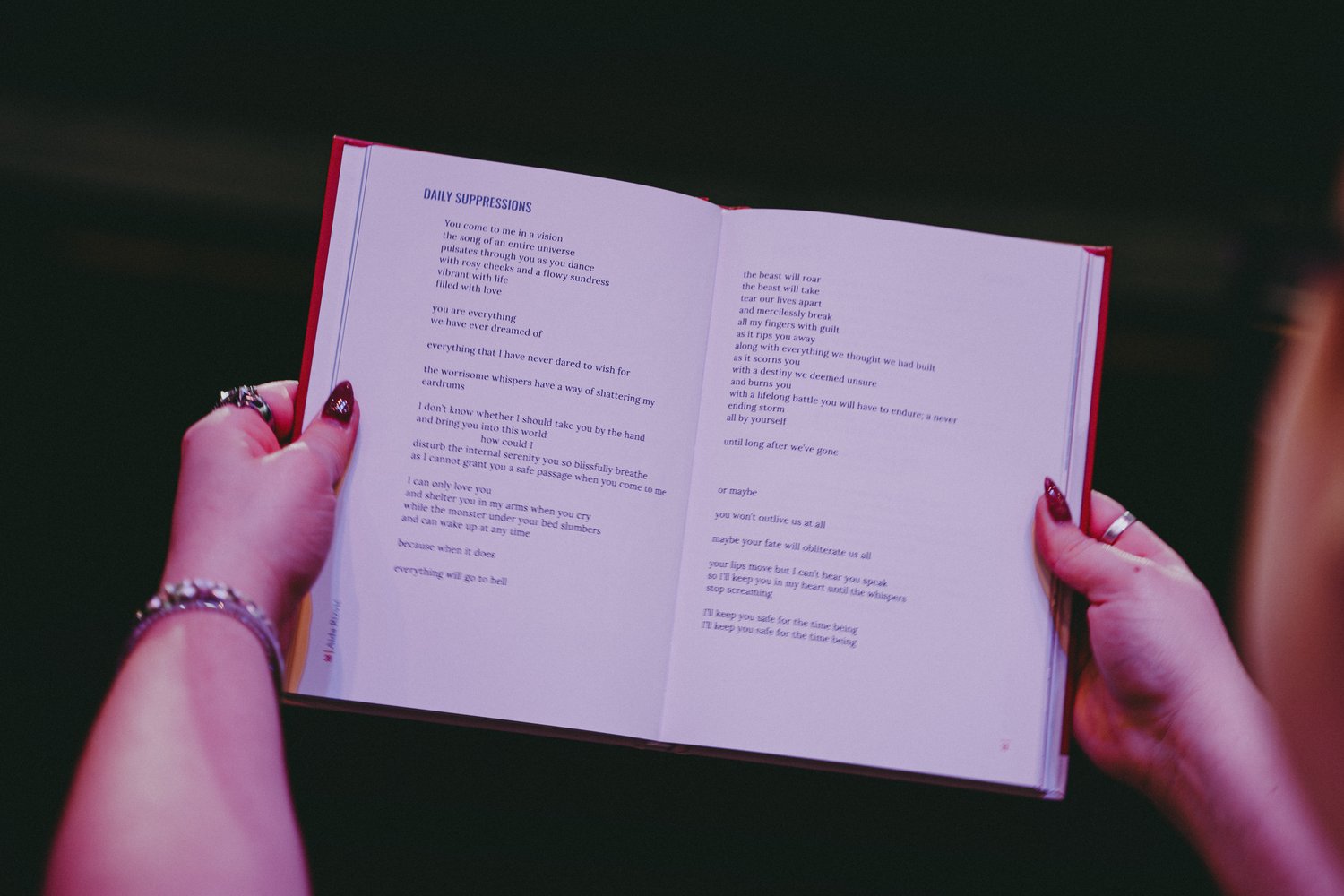 Foto van twee handen die een open boek vasthouden in een theaterzaal. Op de achtergrond zijn felle podiumlichten zichtbaar. Op de pagina's van het boek staat een Engelstalig gedicht in zwarte tekst op een witte pagina.