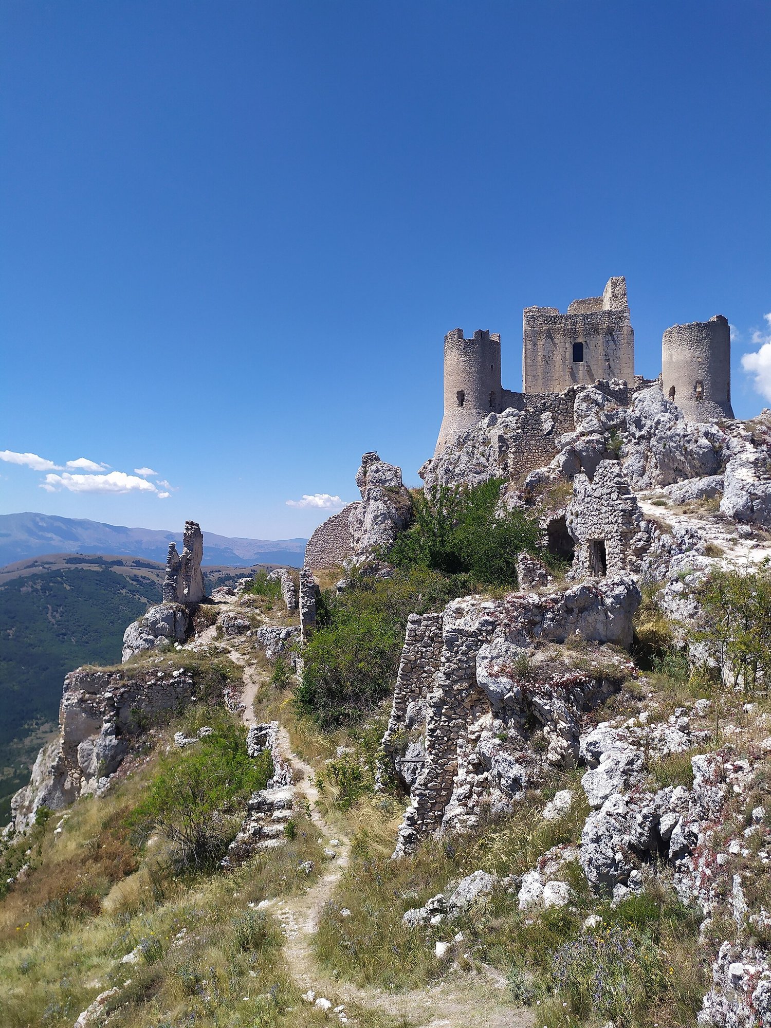 Photograph of a ruined castle on a rocky outcrop. There is a steep and narrow path leading to it and the sky is a deep blue.