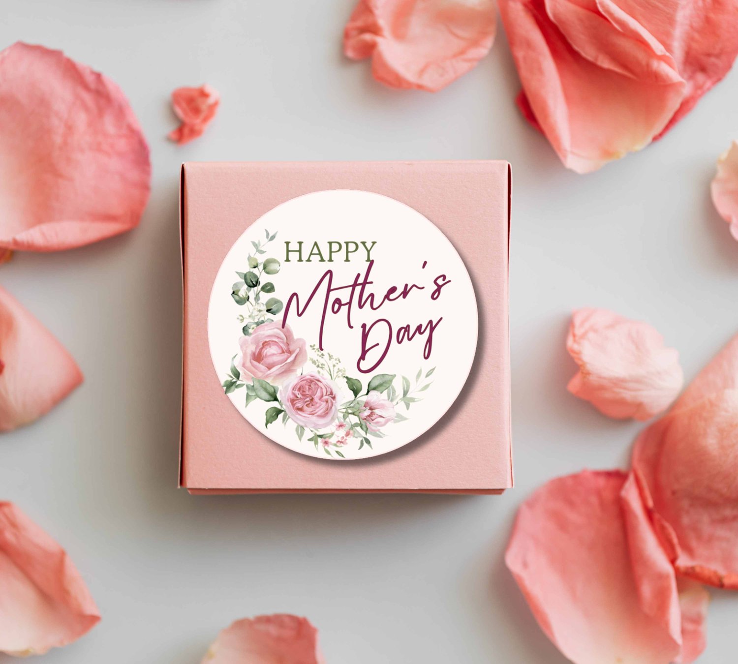 Pink gift box topped with a round floral sticker reading “HAPPY Mother’s Day,” surrounded by scattered rose petals.