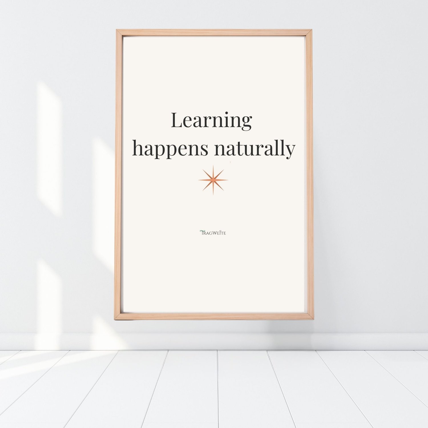 Framed printable poster "Learning happens naturally" with terracotta star, leaning against a white wall on a light wooden floor. Minimalist wall art by Tragweite.  Gerahmtes Poster „Lernen geschieht natürlich" mit terrakottafarbenem Stern, an eine weiße W