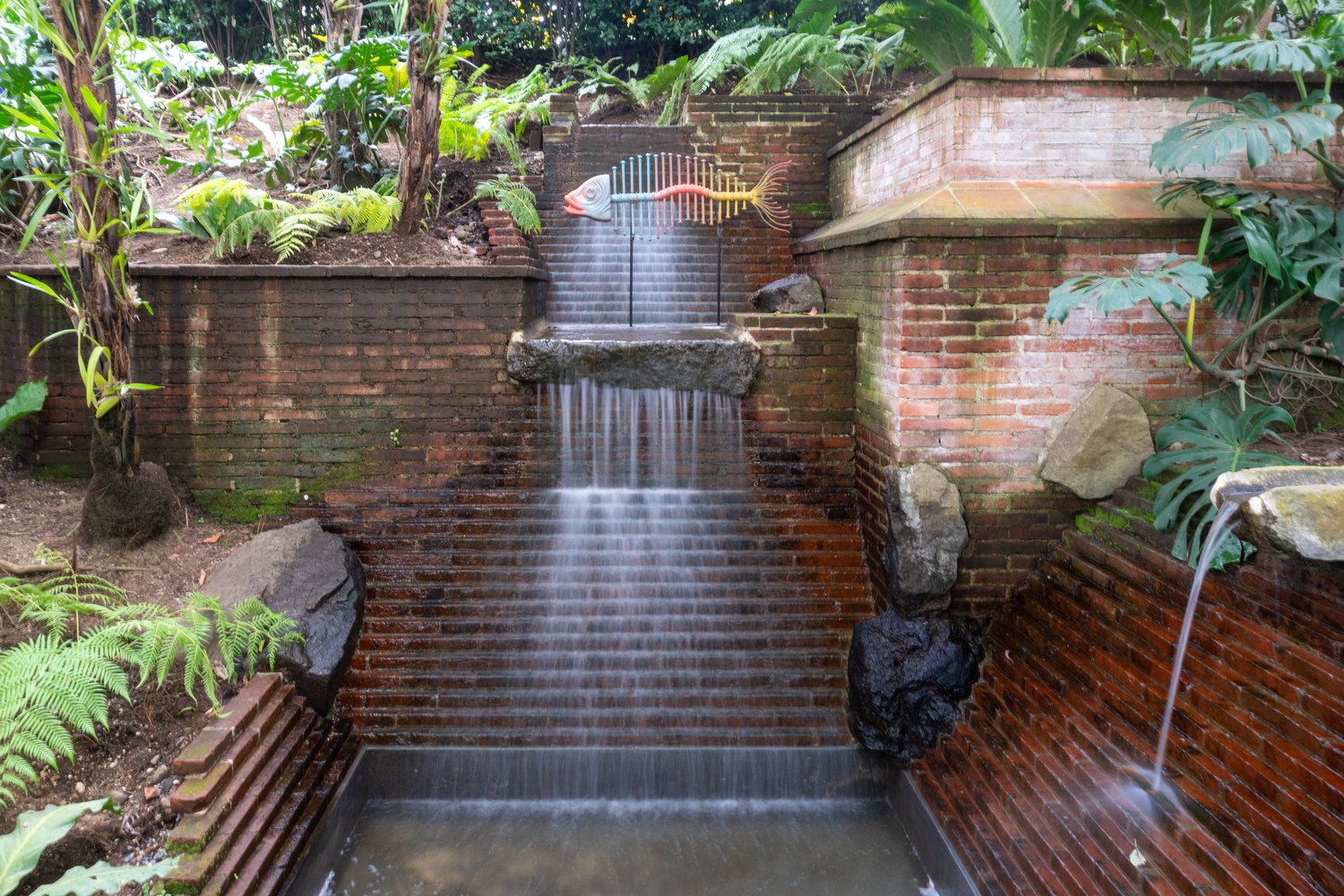 A horizontal long-exposure photograph of a tiered red brick fountain with cascading water, surrounded by tropical green plants and a colorful metal fish sculpture at the top in Antigua.