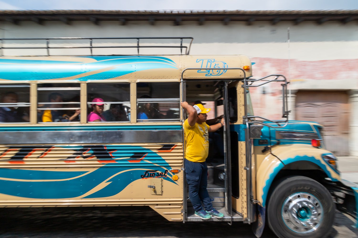 A side-view panning photograph of a brightly decorated cream and blue bus speeding past a colonial wall in Antigua, Guatemala, with a worker in a yellow shirt standing in the open doorway.