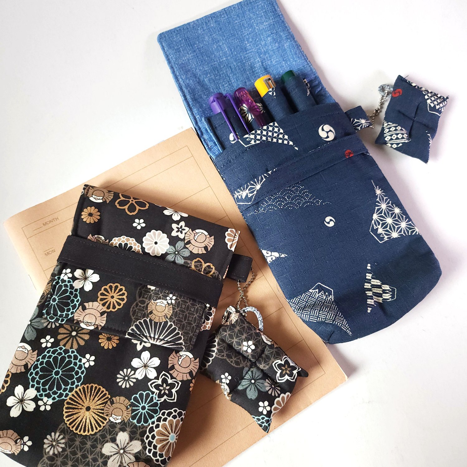 "Top-down view of two handmade fabric pen cases: one in blue Mt. Fuji print and one in black with gold floral patterns, displayed next to a cream-colored notebook."