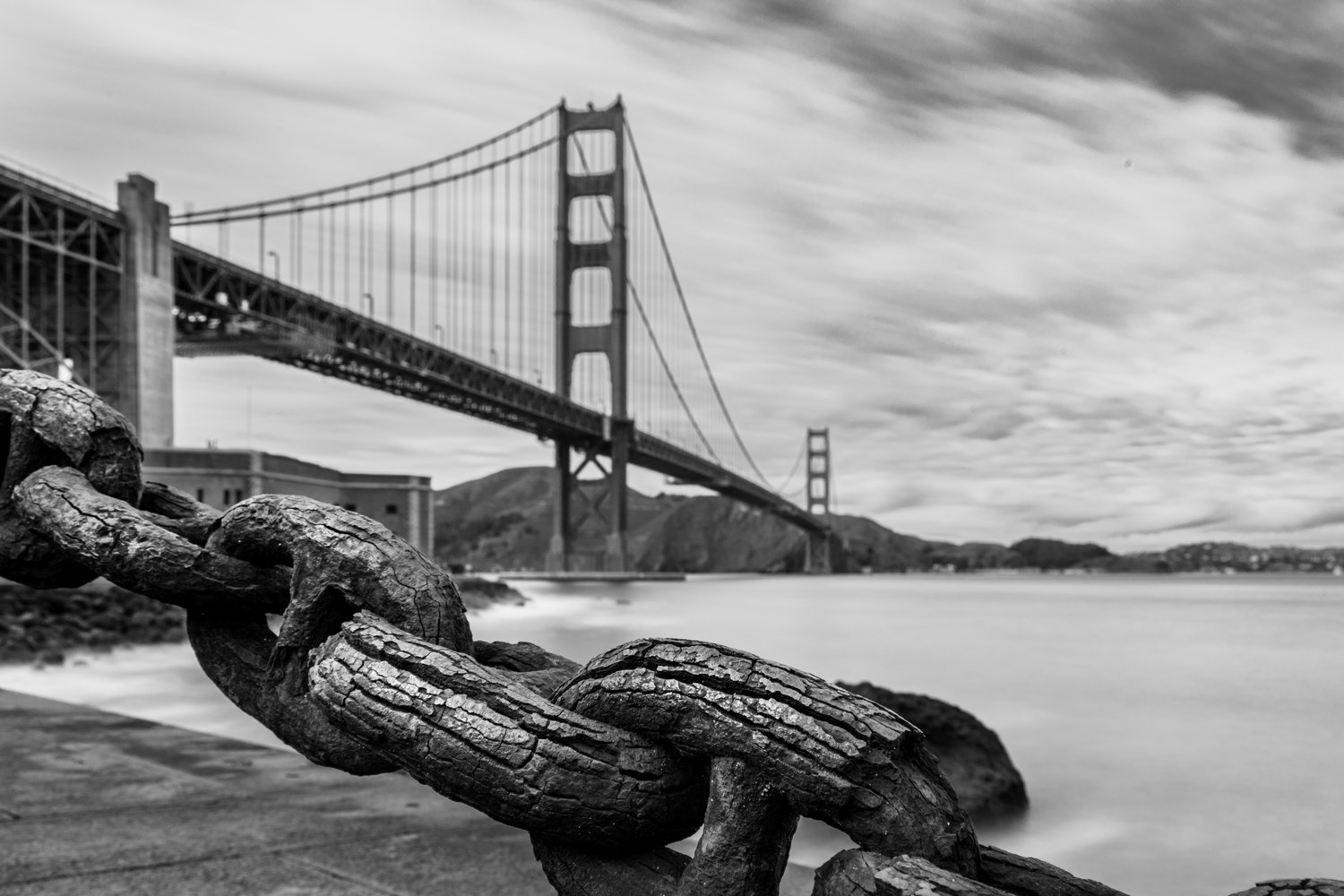 A dramatic black and white long-exposure photograph of the Golden Gate Bridge stretching across smooth water, anchored by large, heavily weathered iron chain links in the sharp foreground.