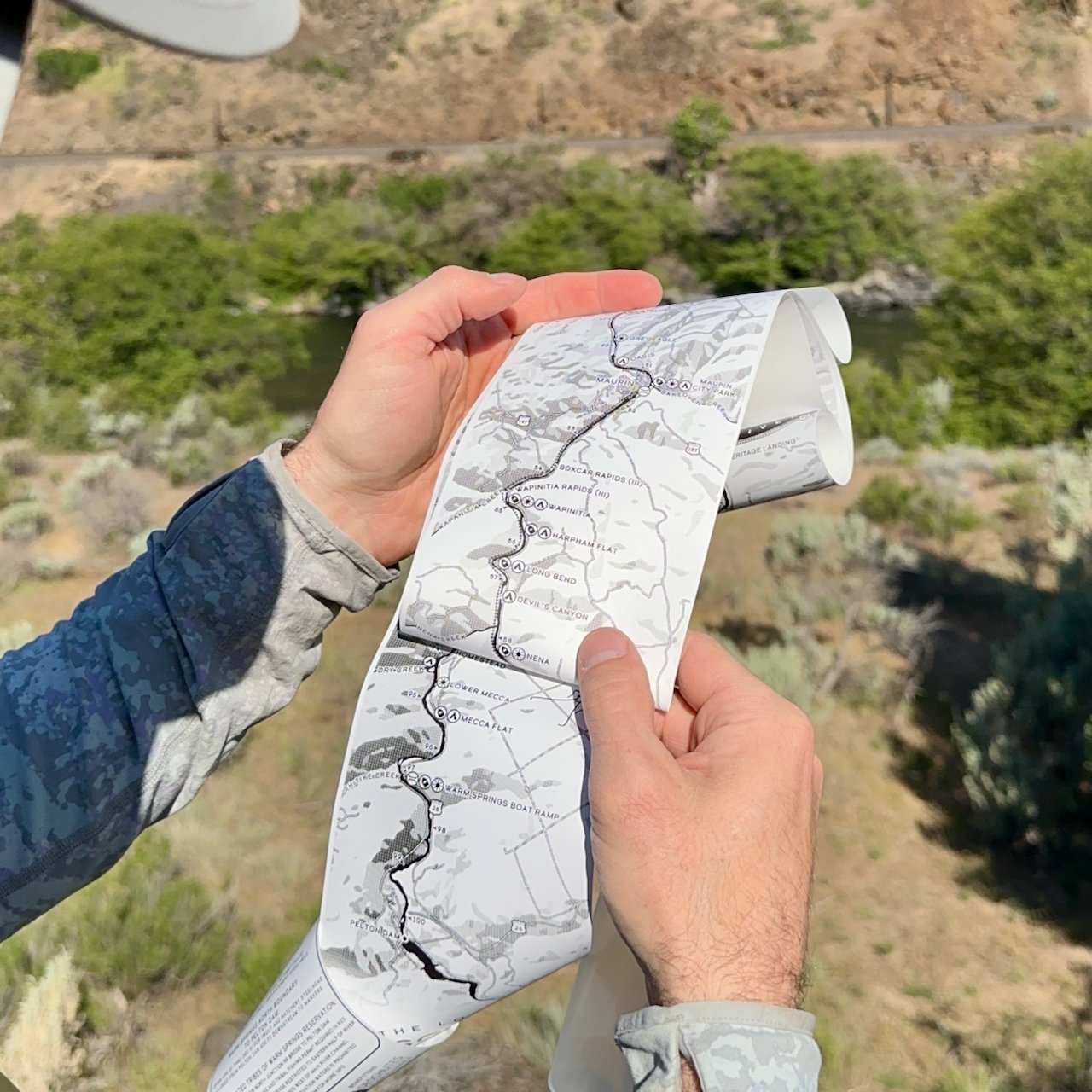 Angler holding the Lower Deschutes Ribbon Map open above the river canyon, showing campsites and rapids from Maupin downstream, with sagebrush hillsides and the Deschutes River visible below