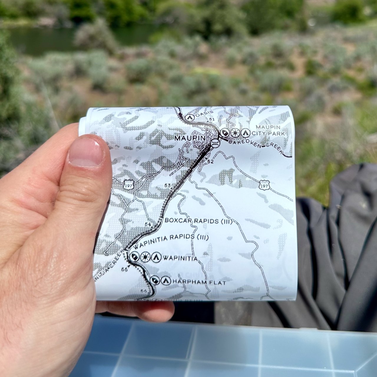 Lower Deschutes Ribbon Map rolled in hand at the river, showing Maupin, Boxcar Rapids, Wapinitia Rapids, and Harpham Flat resting on a fly box, with the Deschutes River in the background