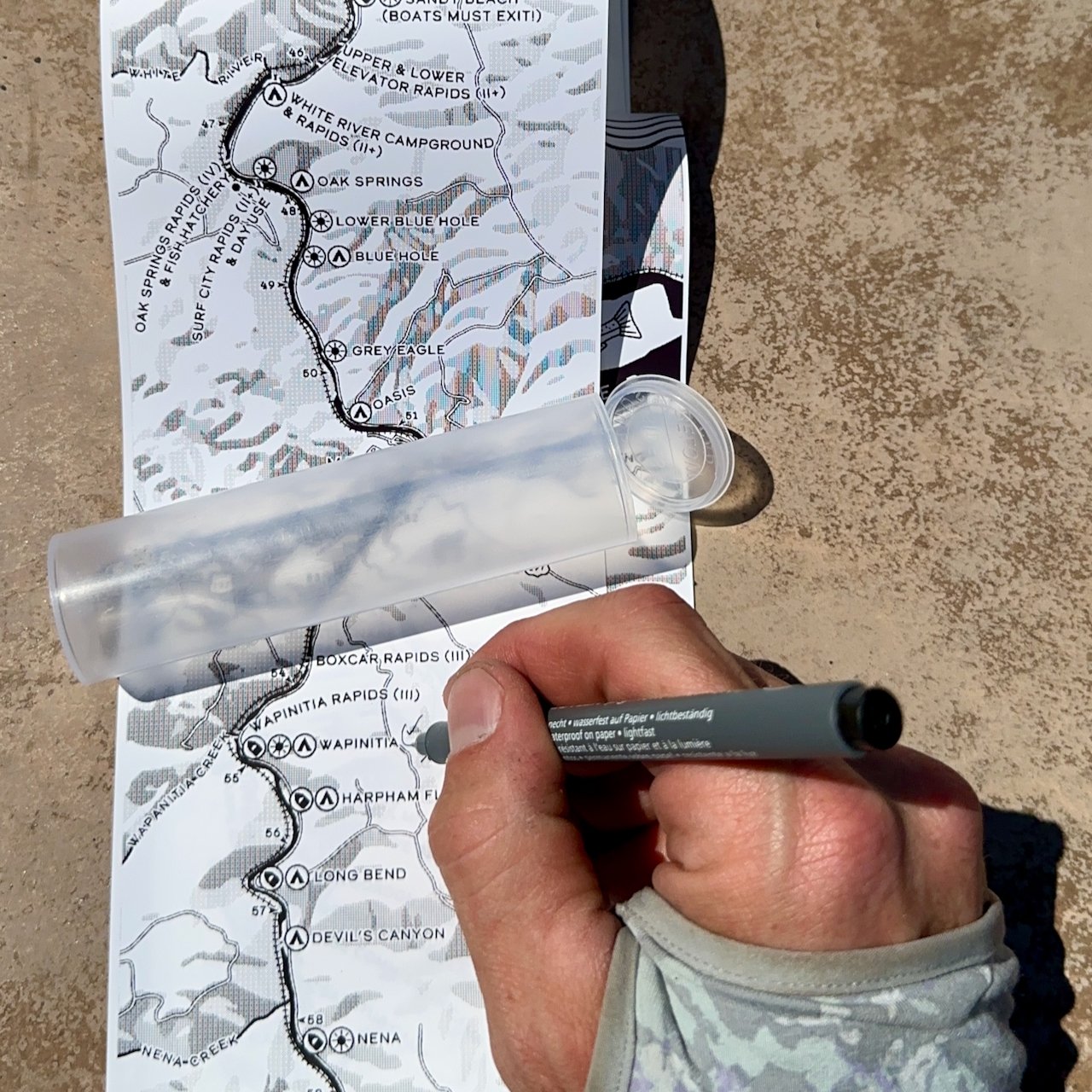 Hand marking the Lower Deschutes Ribbon Map with a waterproof pen at a riverside picnic table, with the clear doob tube storage case visible alongside